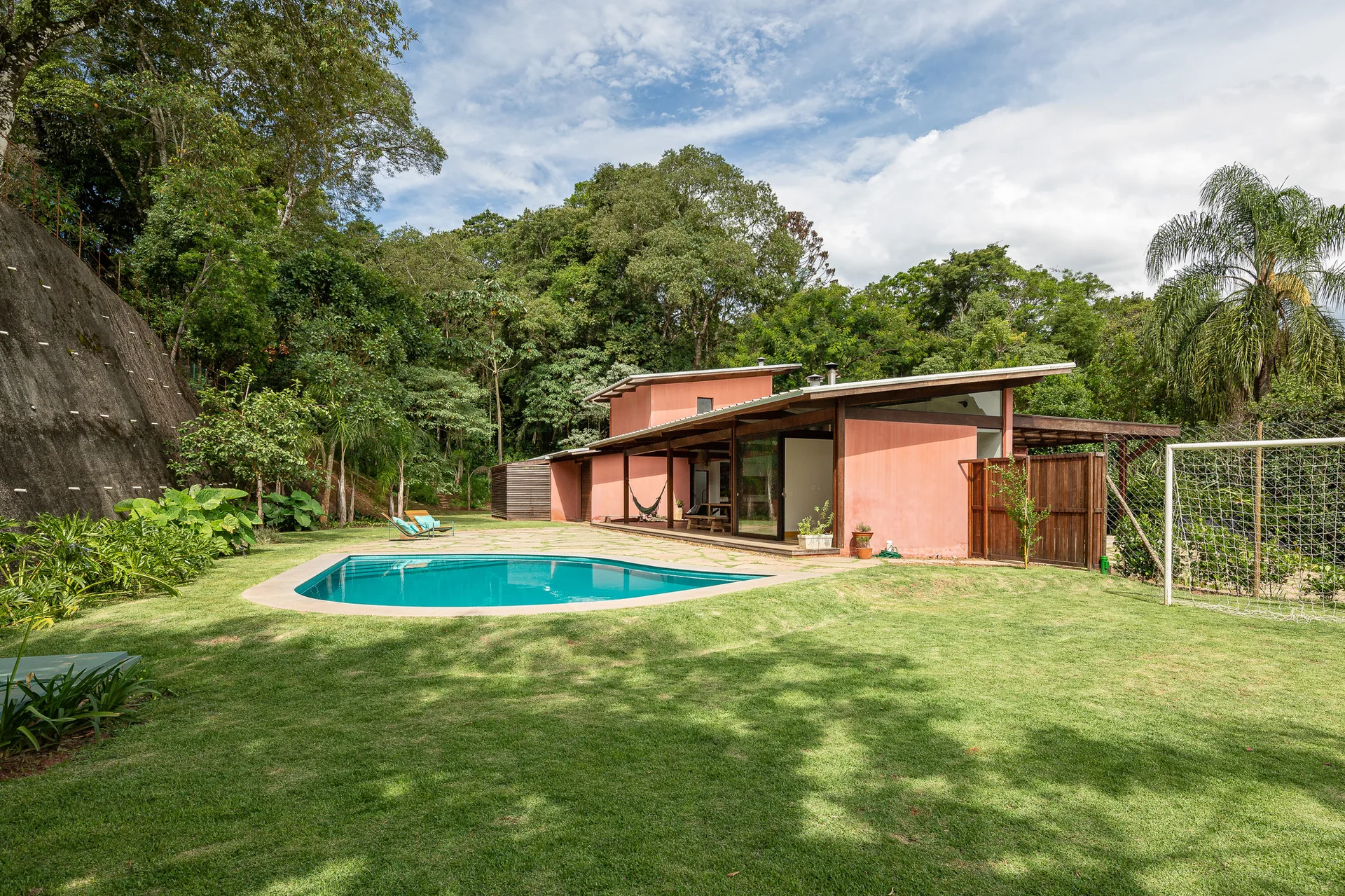A contemporary terracotta house with a shed roof sits next to a kidney-shaped swimming pool on a grassy lawn surrounded by dense trees.