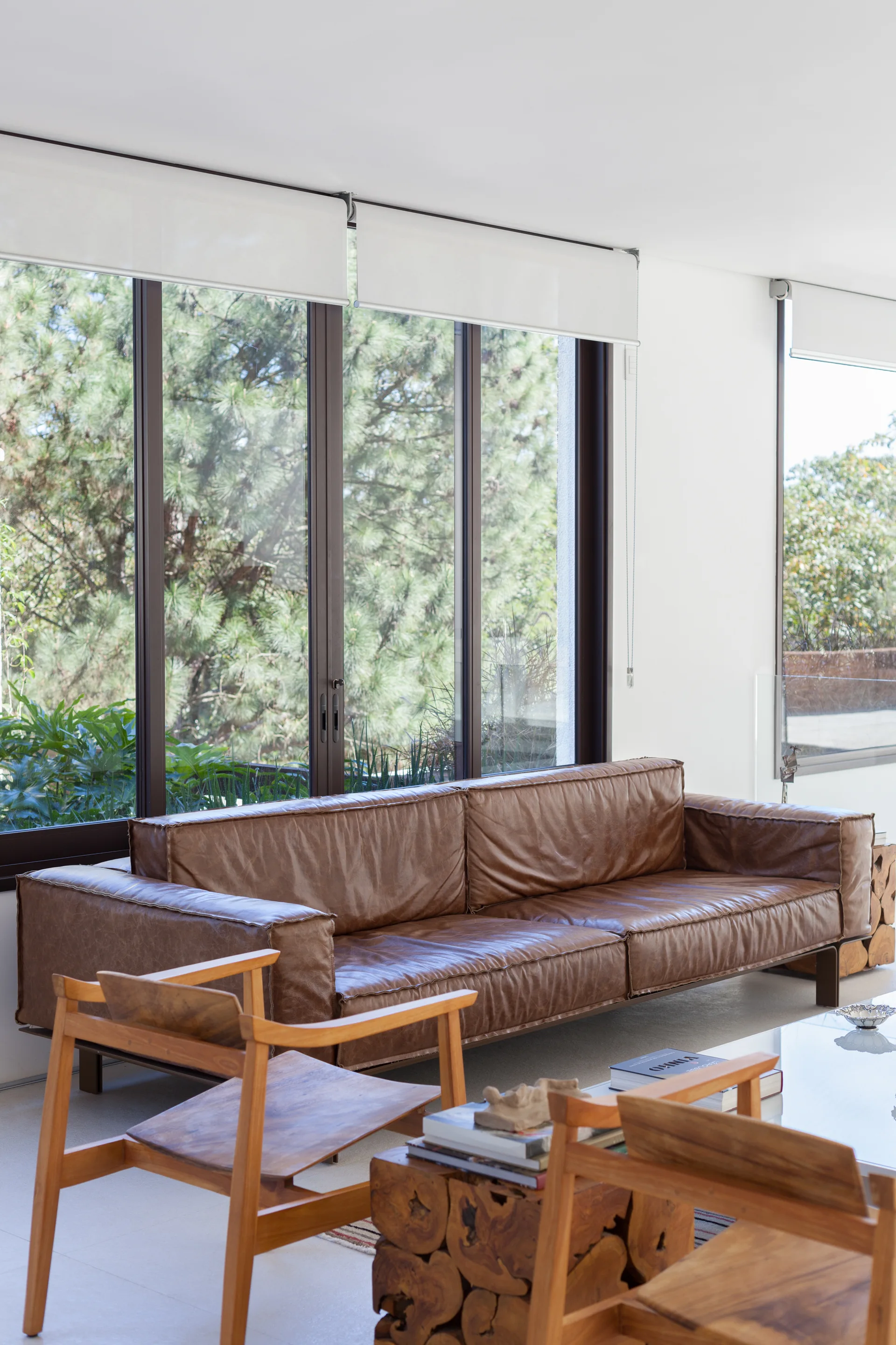 A modern white living room with a low brown leather sofa, wood armchairs, and a block coffee table in front of large windows.