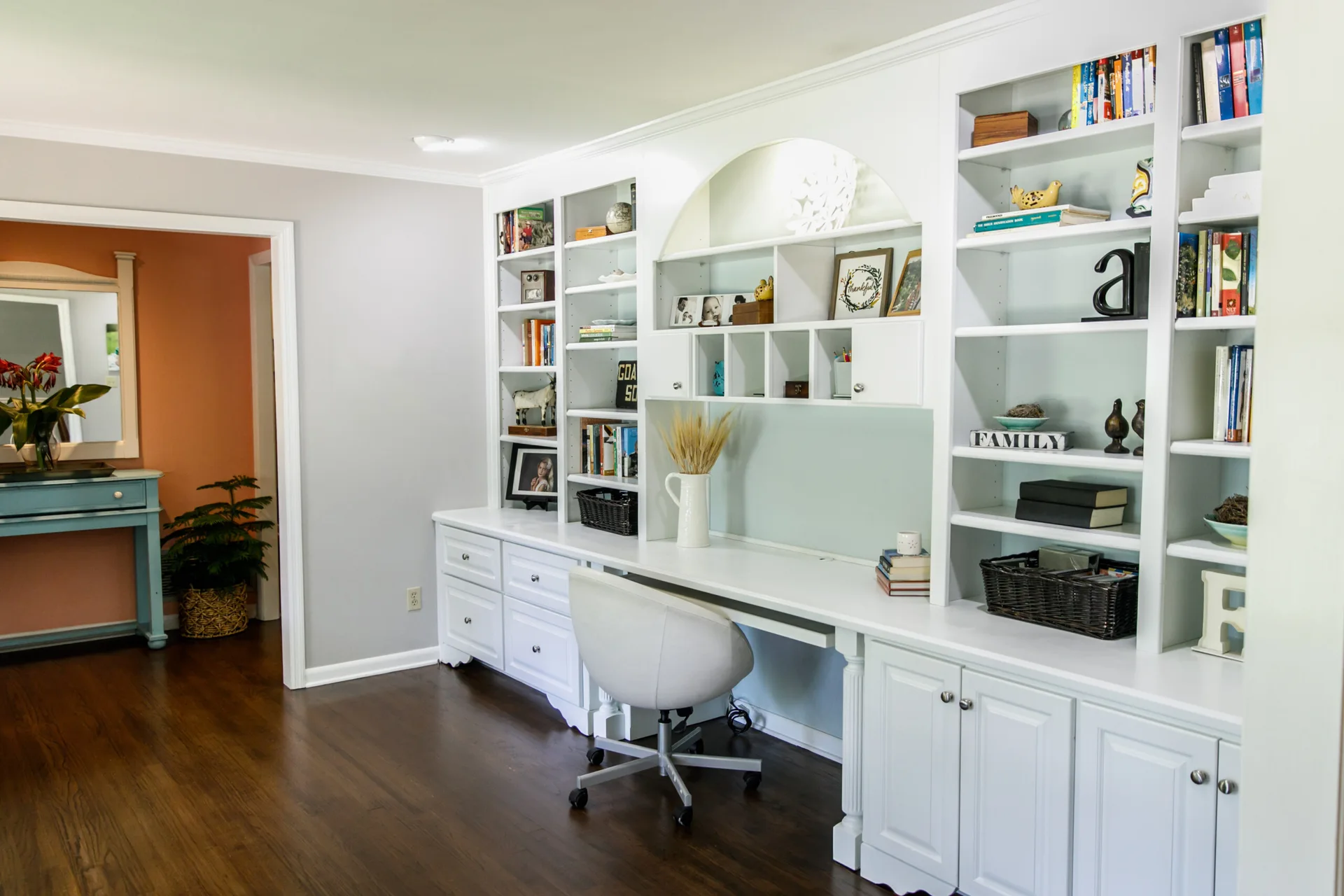 A home office features a large white built-in desk with bookshelves, a pale blue backing, and a white office chair on dark wood floors.