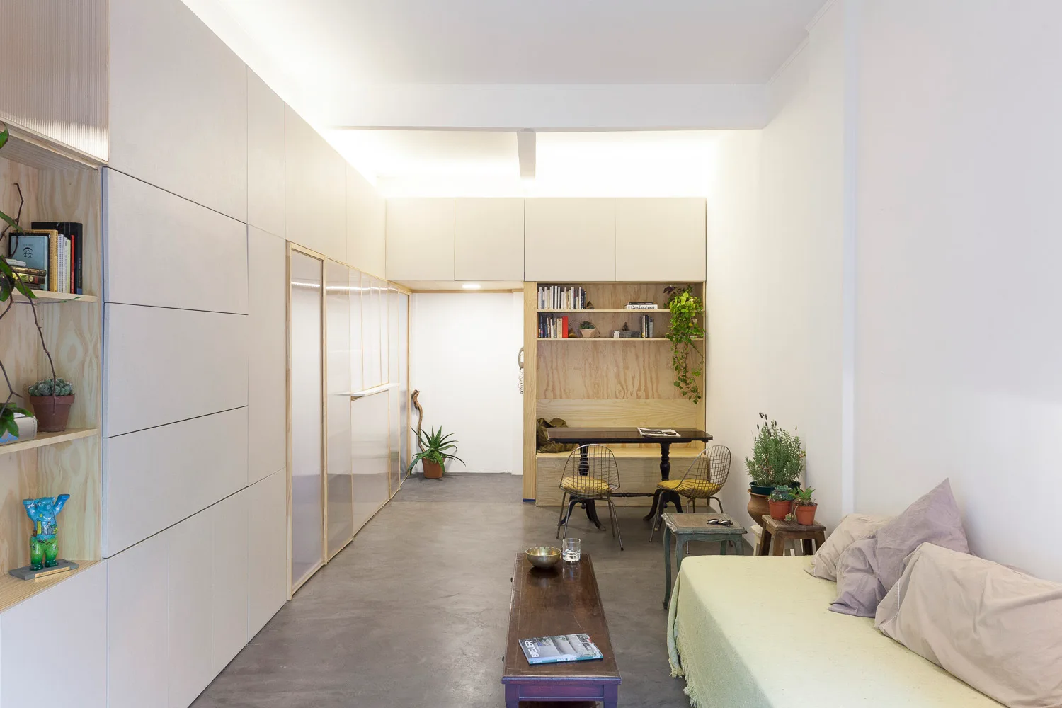 A minimalist living room features flat-panel storage cabinets, a built-in plywood dining nook, concrete flooring, and a simple green daybed.