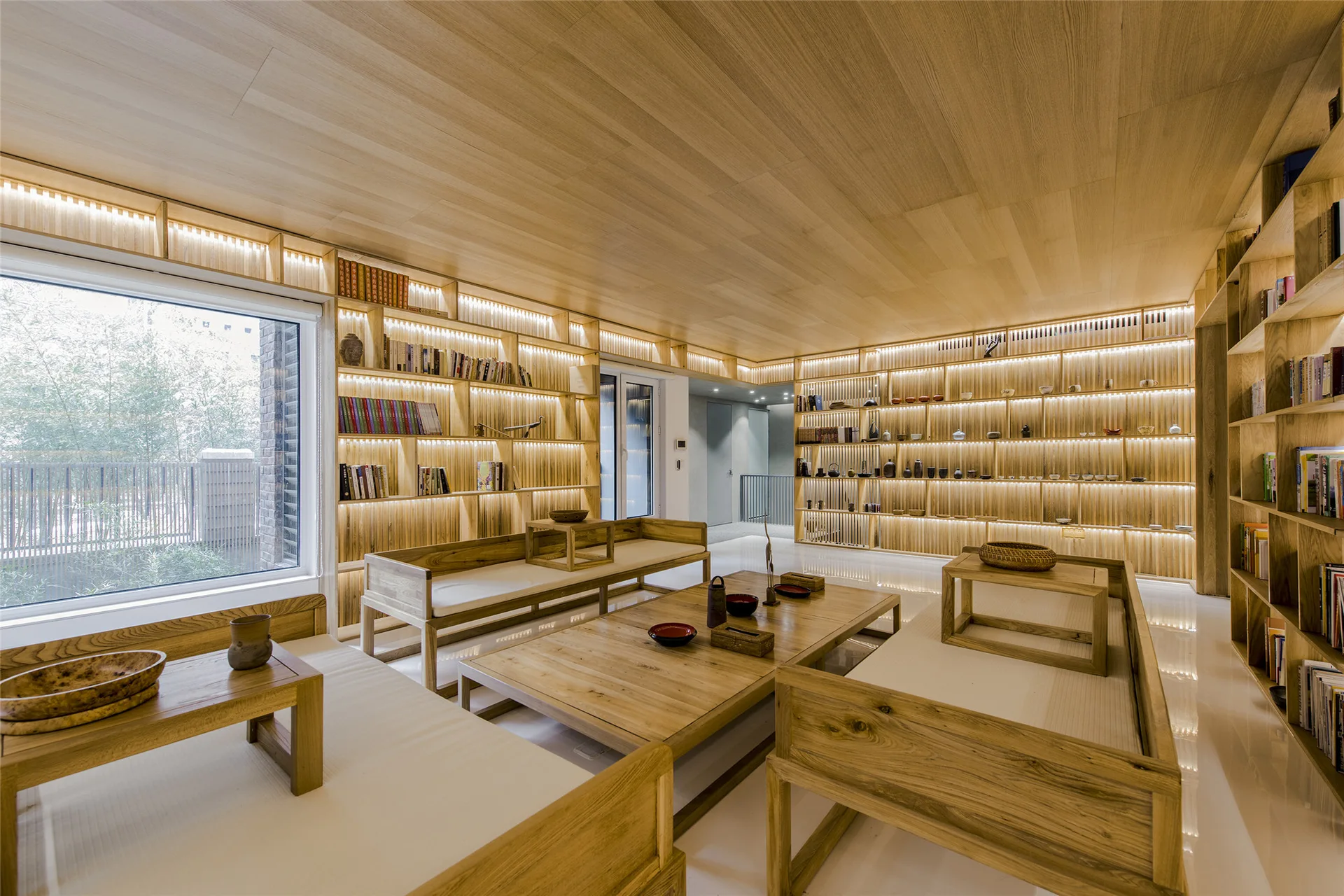 A modern lounge features extensive light wood wall shelving with integrated LED lighting, matching low-profile furniture, and a glossy white floor.