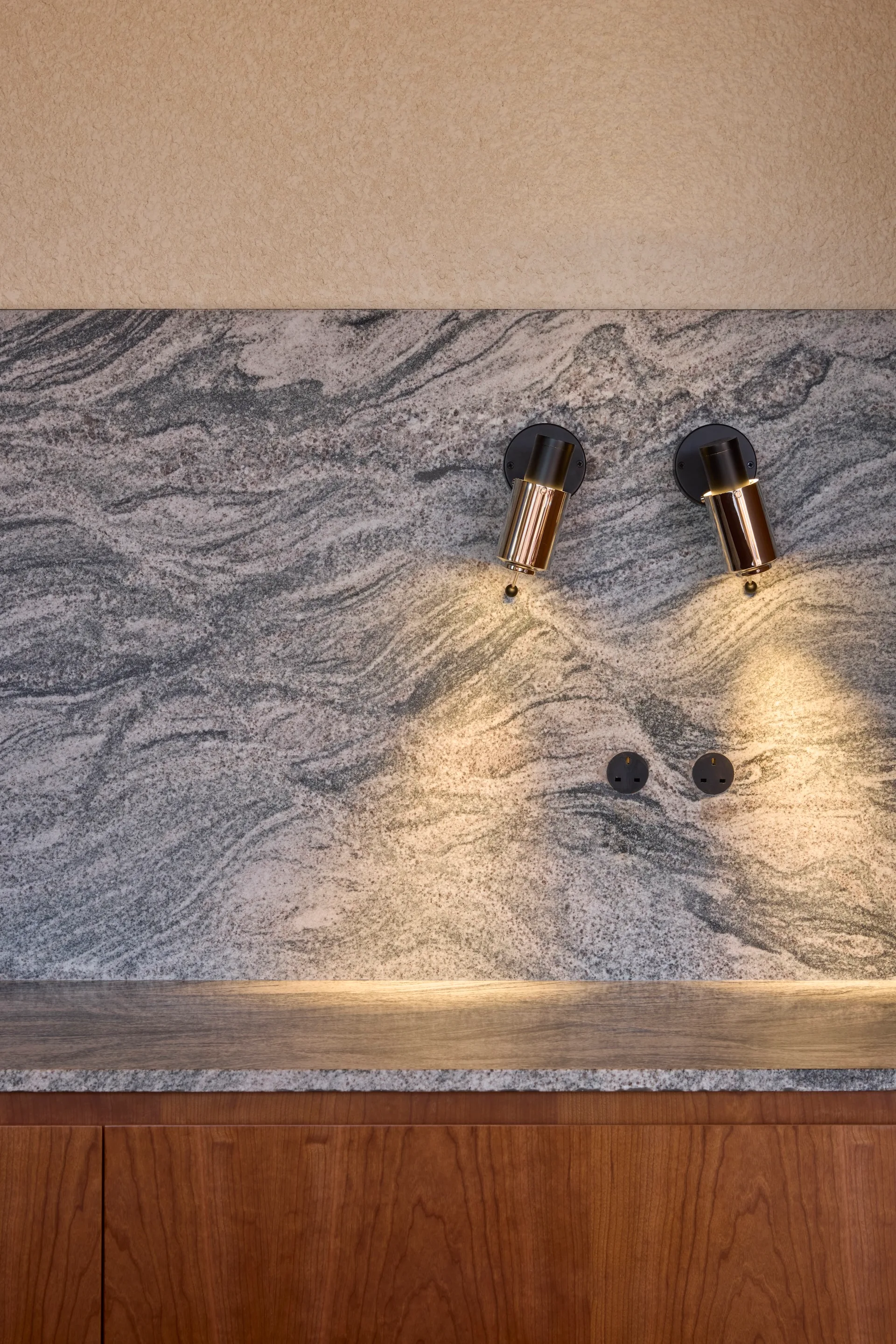 A grey veined stone backsplash features two cylindrical brass wall sconces above warm wood kitchen cabinetry.