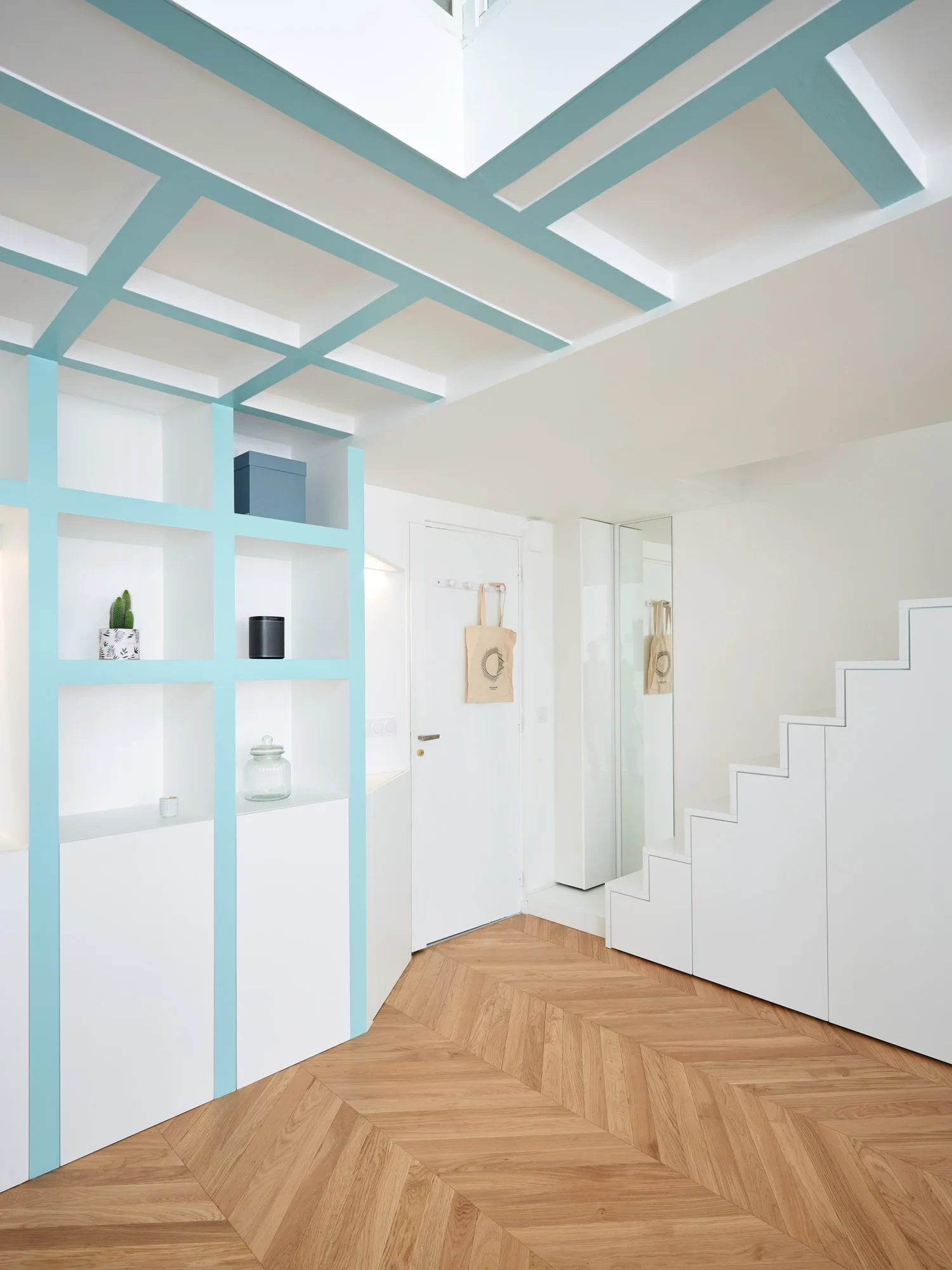 A modern entryway with white custom built-in shelves featuring teal accents, a matching ceiling grid, storage under the stairs, and light wood chevron floors.