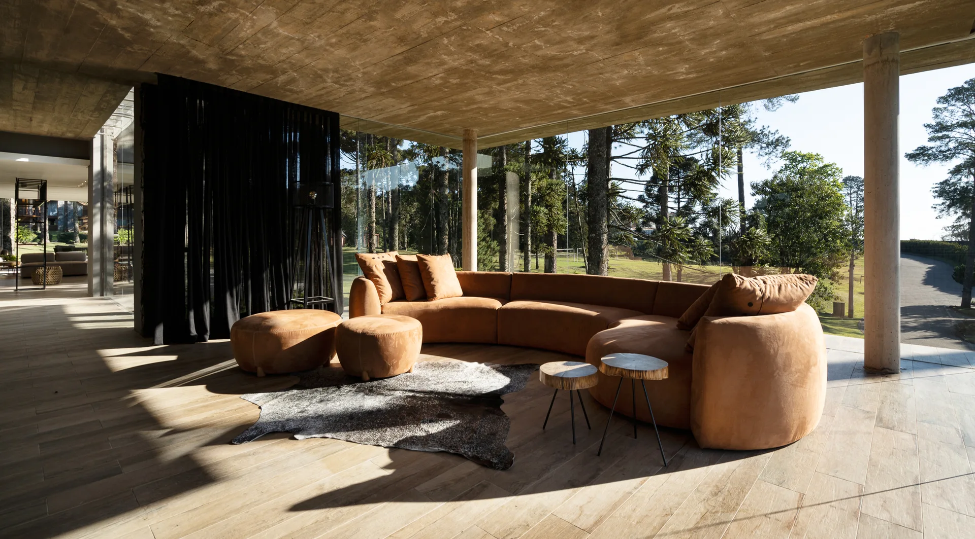 A curved tan sofa and matching ottomans on an animal hide rug in a living room with floor-to-ceiling glass walls and a raw concrete ceiling.