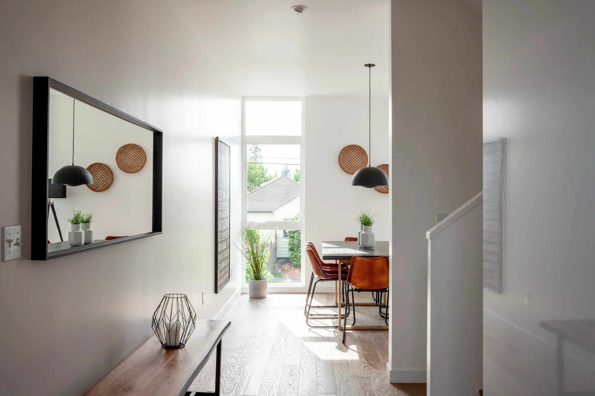 A bright hallway with a wooden bench and large mirror leading into a sunlit dining room with leather chairs and a black pendant light.
