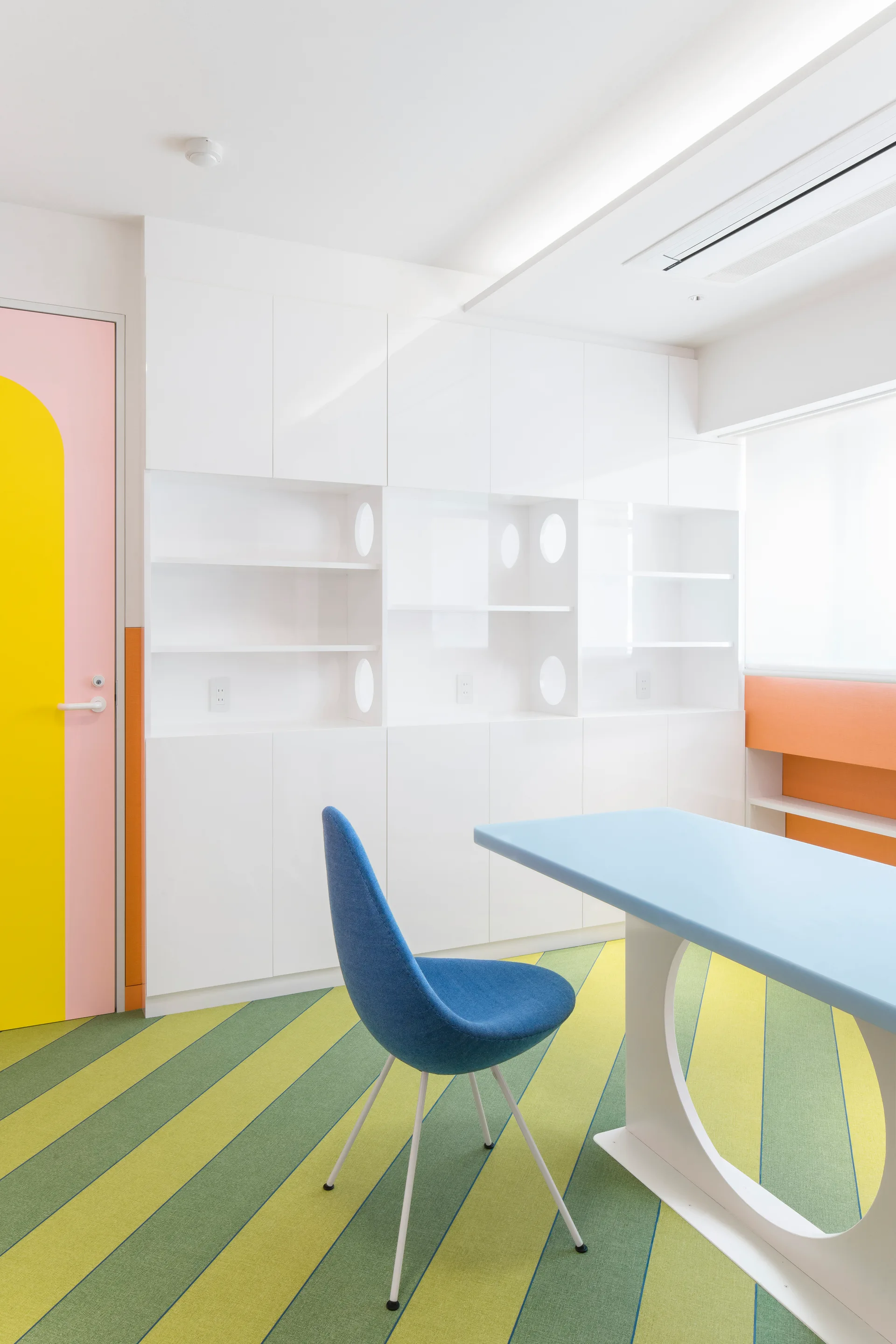 A modern white workspace featuring built-in cabinetry, a light blue desk with a blue chair, and a diagonal green and yellow striped carpet.