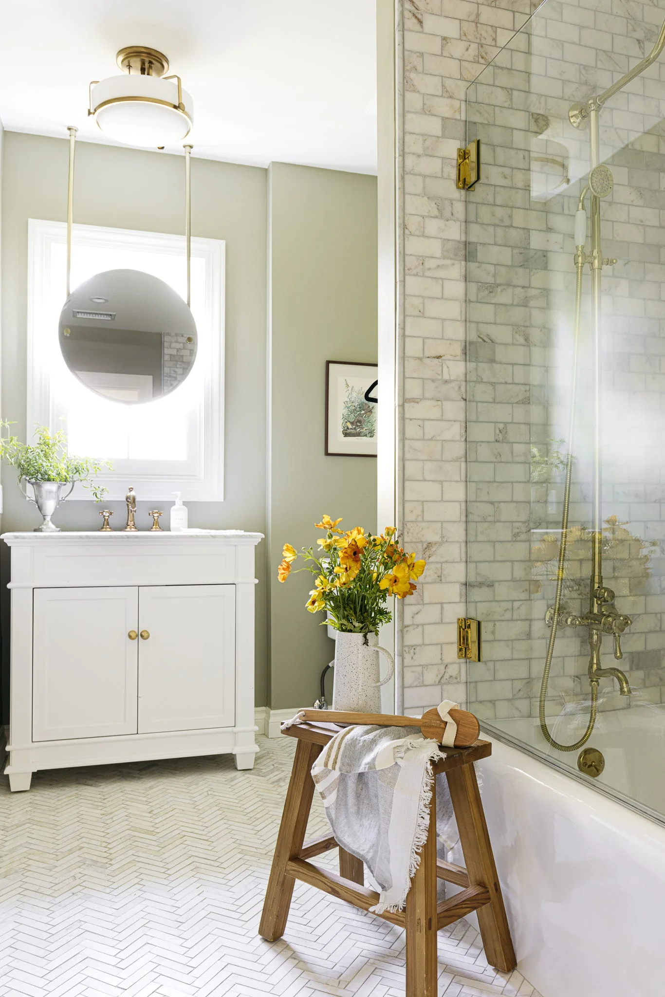 A sage green bathroom features a white vanity, a round suspended mirror in front of a window, and a glass-enclosed shower with brass fixtures.
