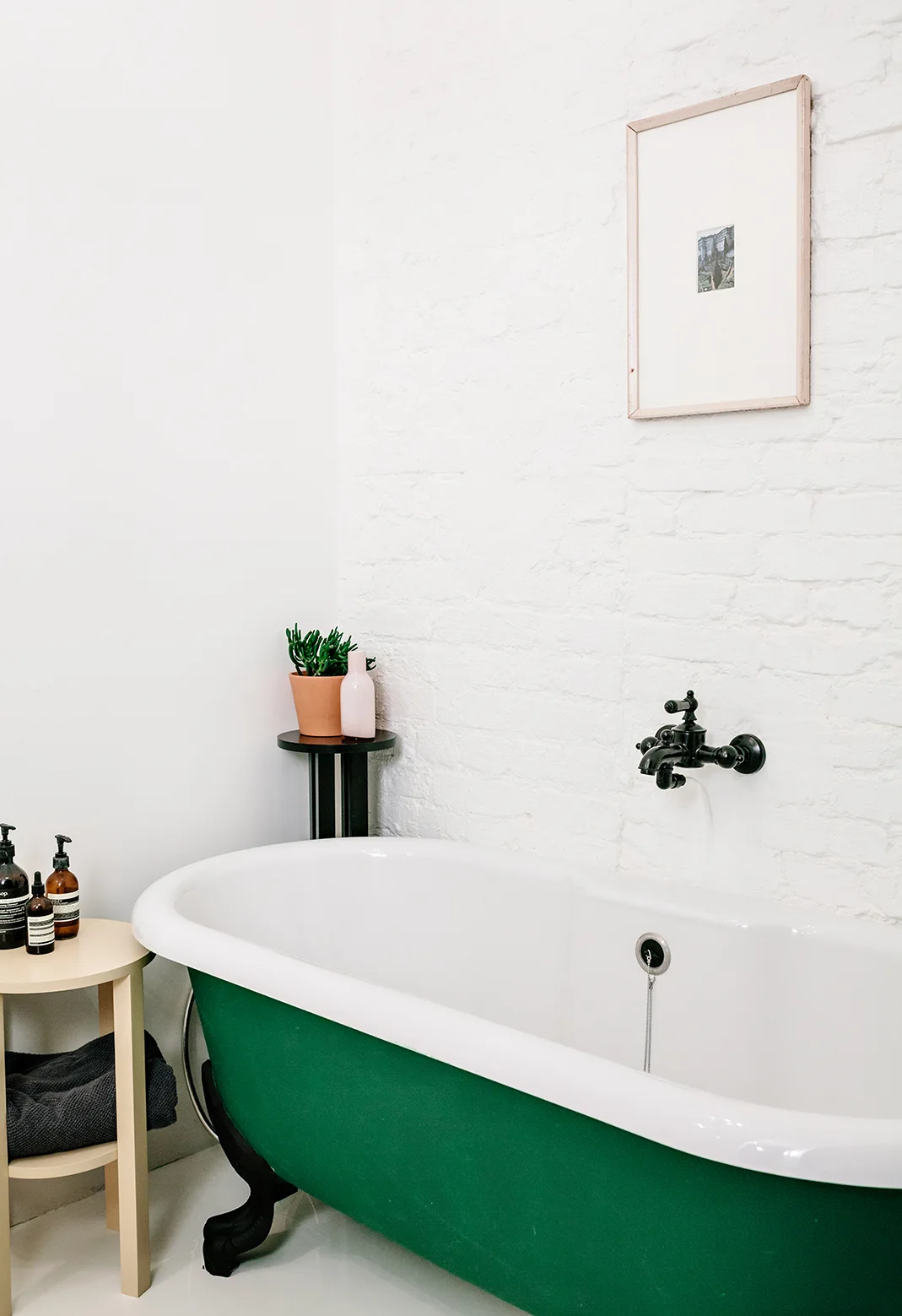 A green clawfoot bathtub sits against a white painted brick wall with a matte black wall-mounted faucet and minimalist side tables.