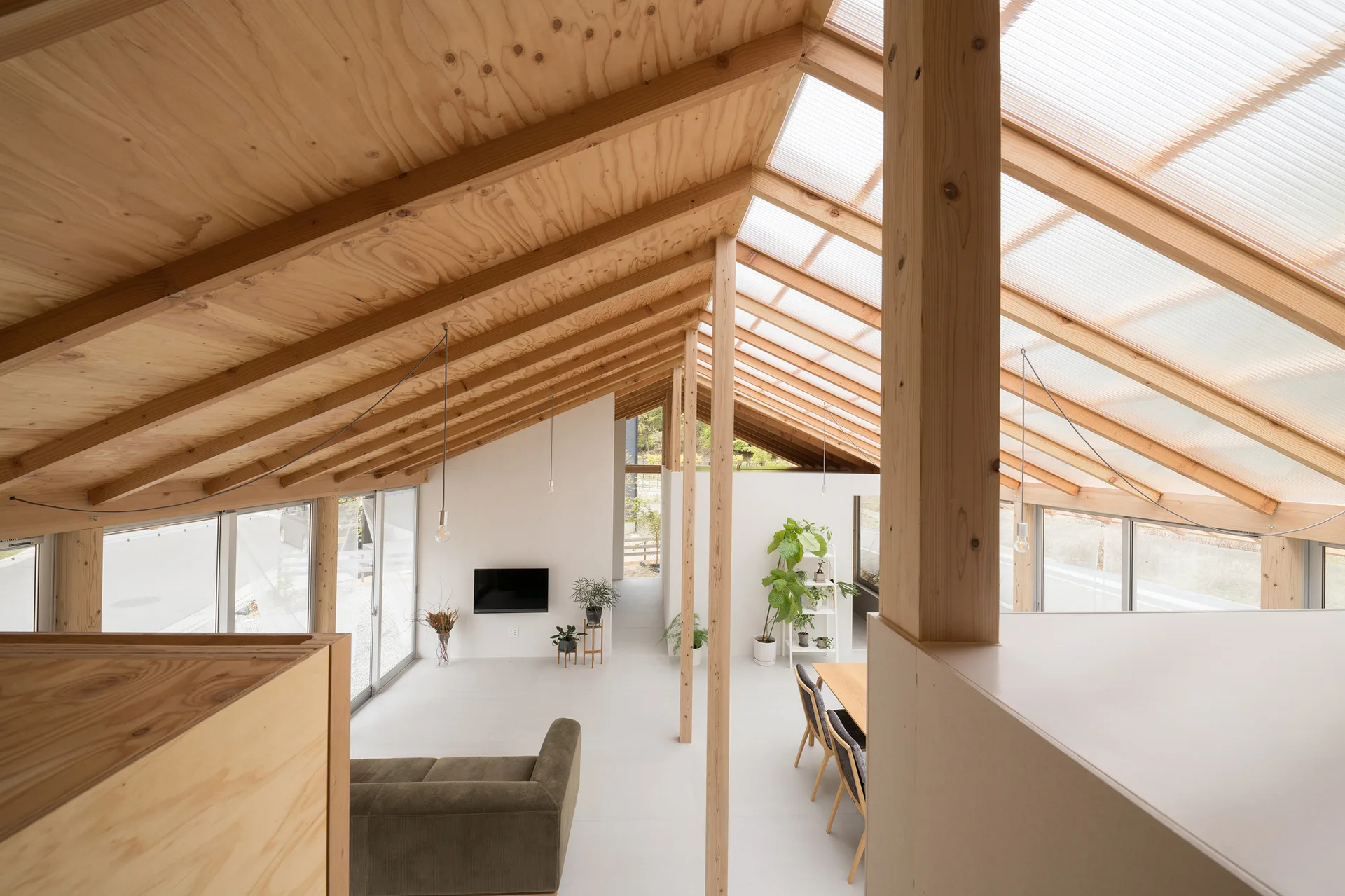 An open-concept living area with a pitched roof featuring exposed plywood rafters, translucent skylight panels, white walls, and minimalist furniture.