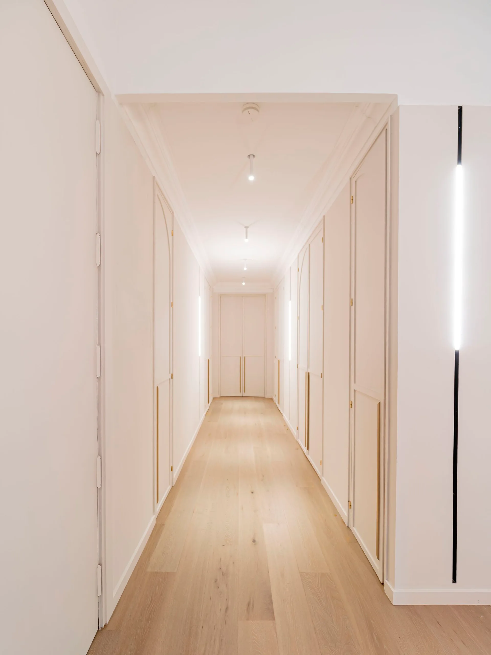 A long white hallway with light wood flooring, floor-to-ceiling built-in closets with gold hardware, and built-in vertical LED light strips.