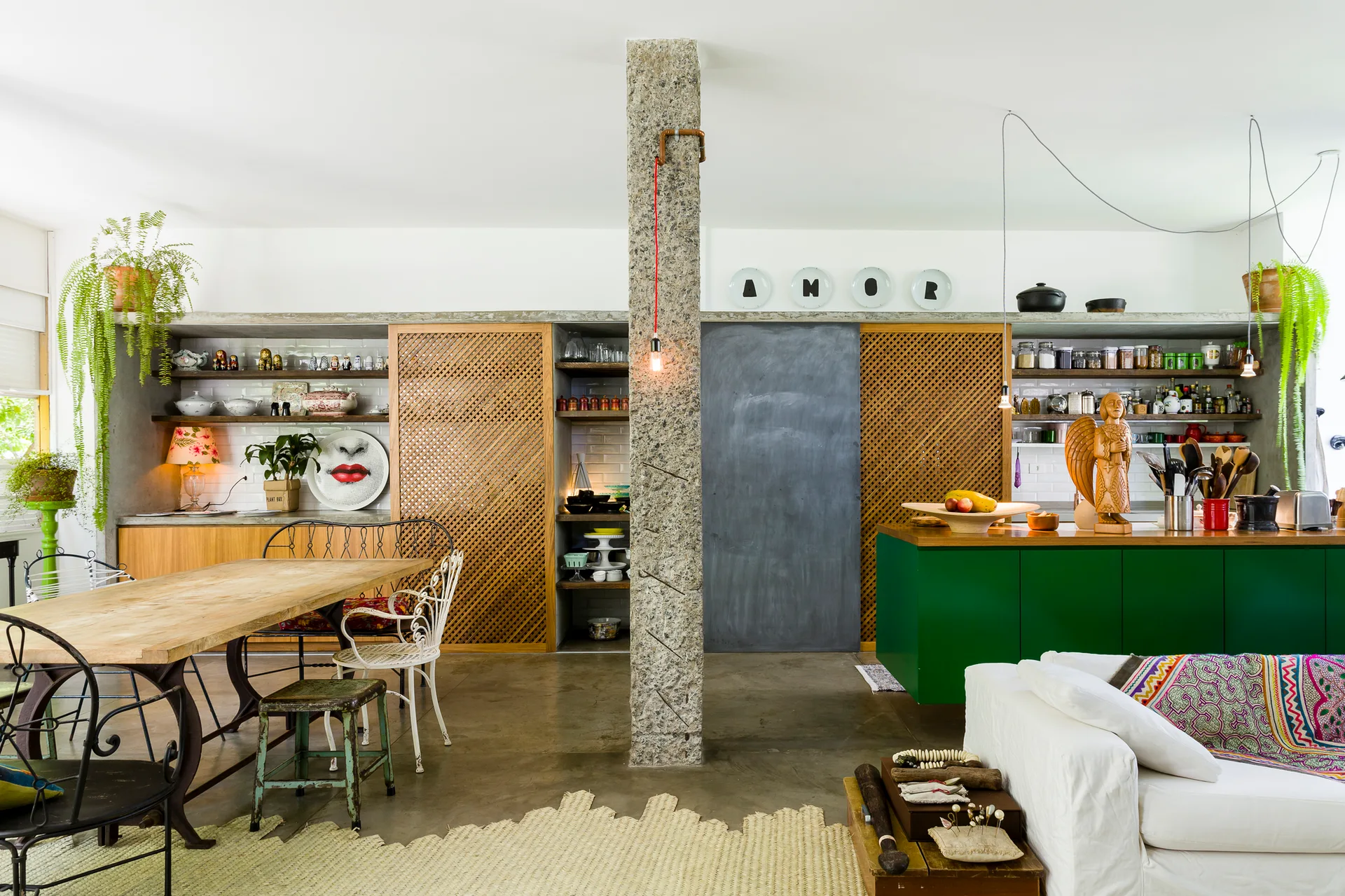 An open-plan eclectic kitchen and dining room features a central stone column, green cabinets, sliding wood-lattice doors, and a rustic wooden dining table.
