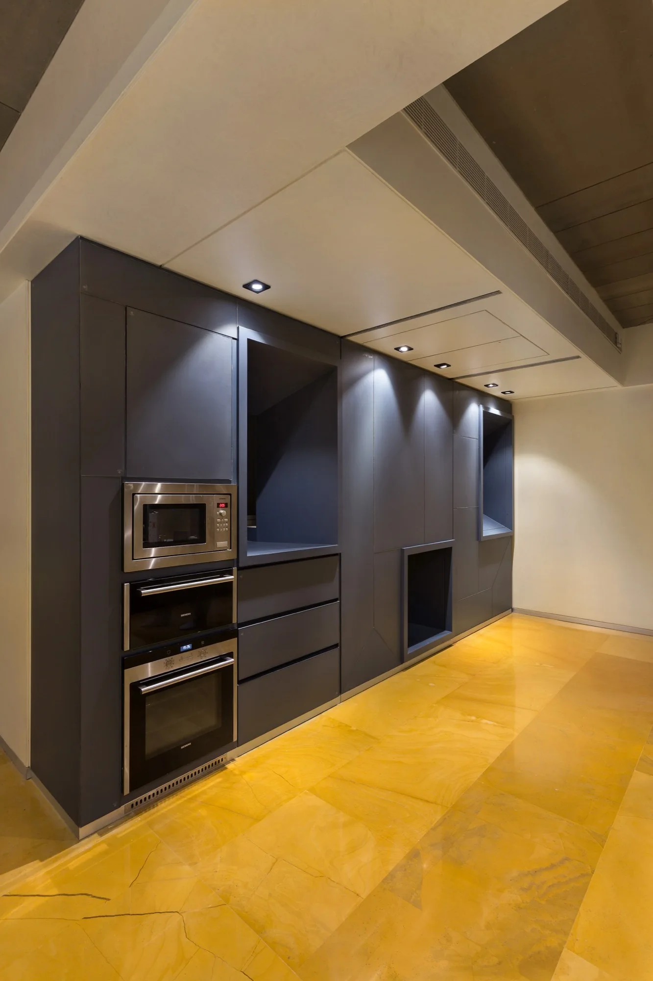A minimalist kitchen featuring dark grey integrated cabinetry with built-in stainless steel ovens and a polished yellow stone floor.