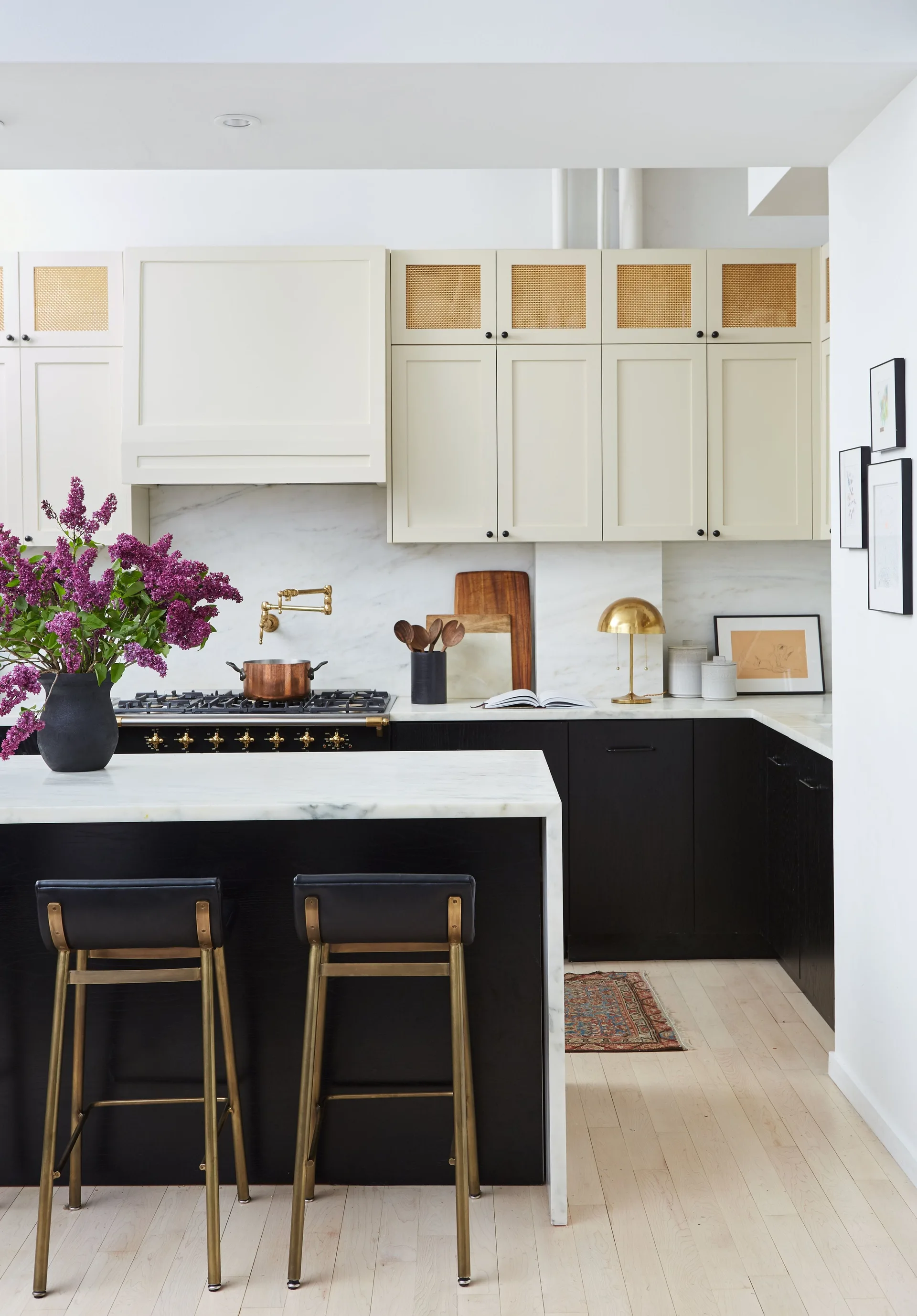 A modern kitchen featuring cream upper cabinets with rattan inserts, black lower cabinets, a marble backsplash, and a black island with brass stools.