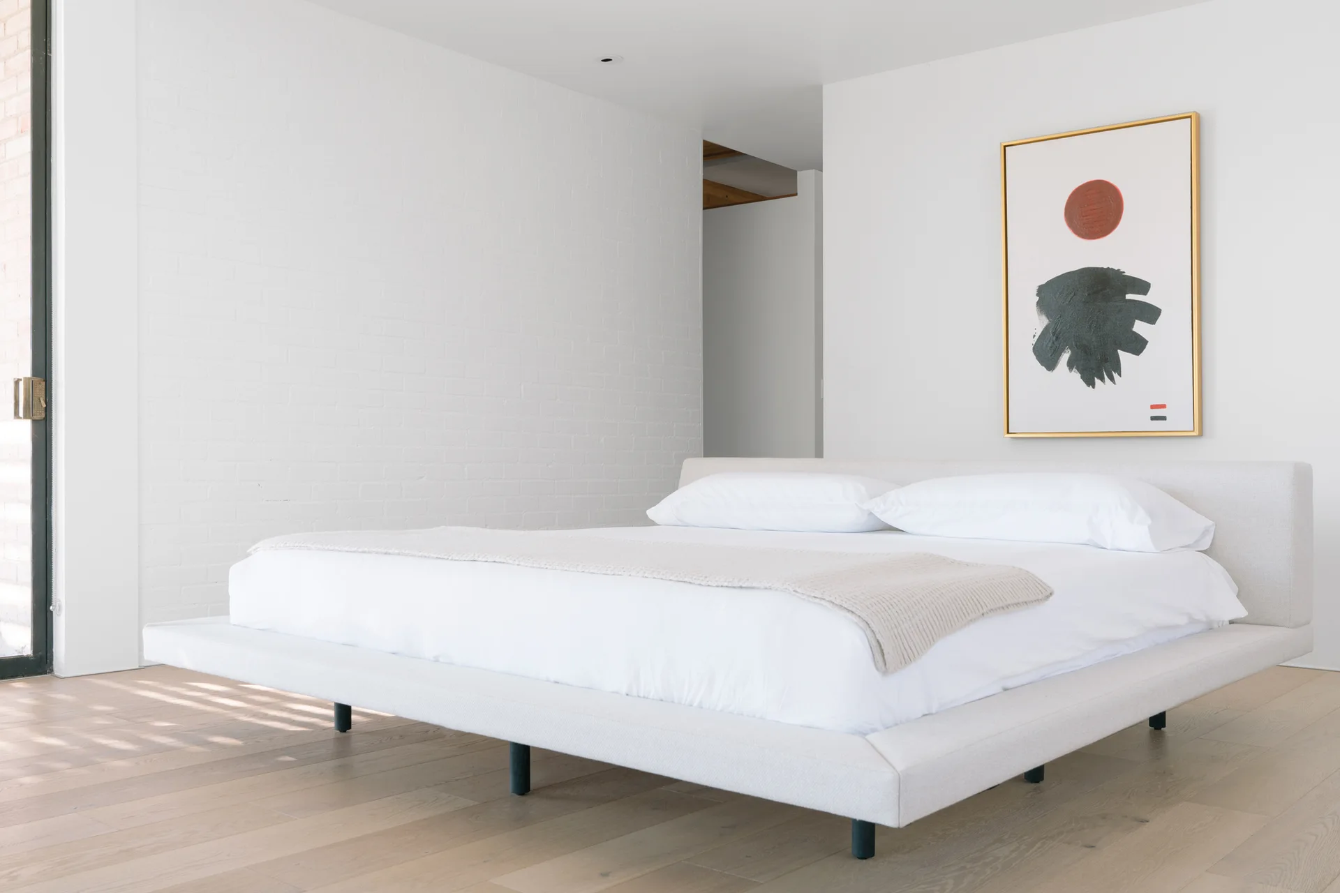 A low-profile upholstered platform bed with white bedding sits on a light oak floor against a white painted brick wall with minimalist artwork.
