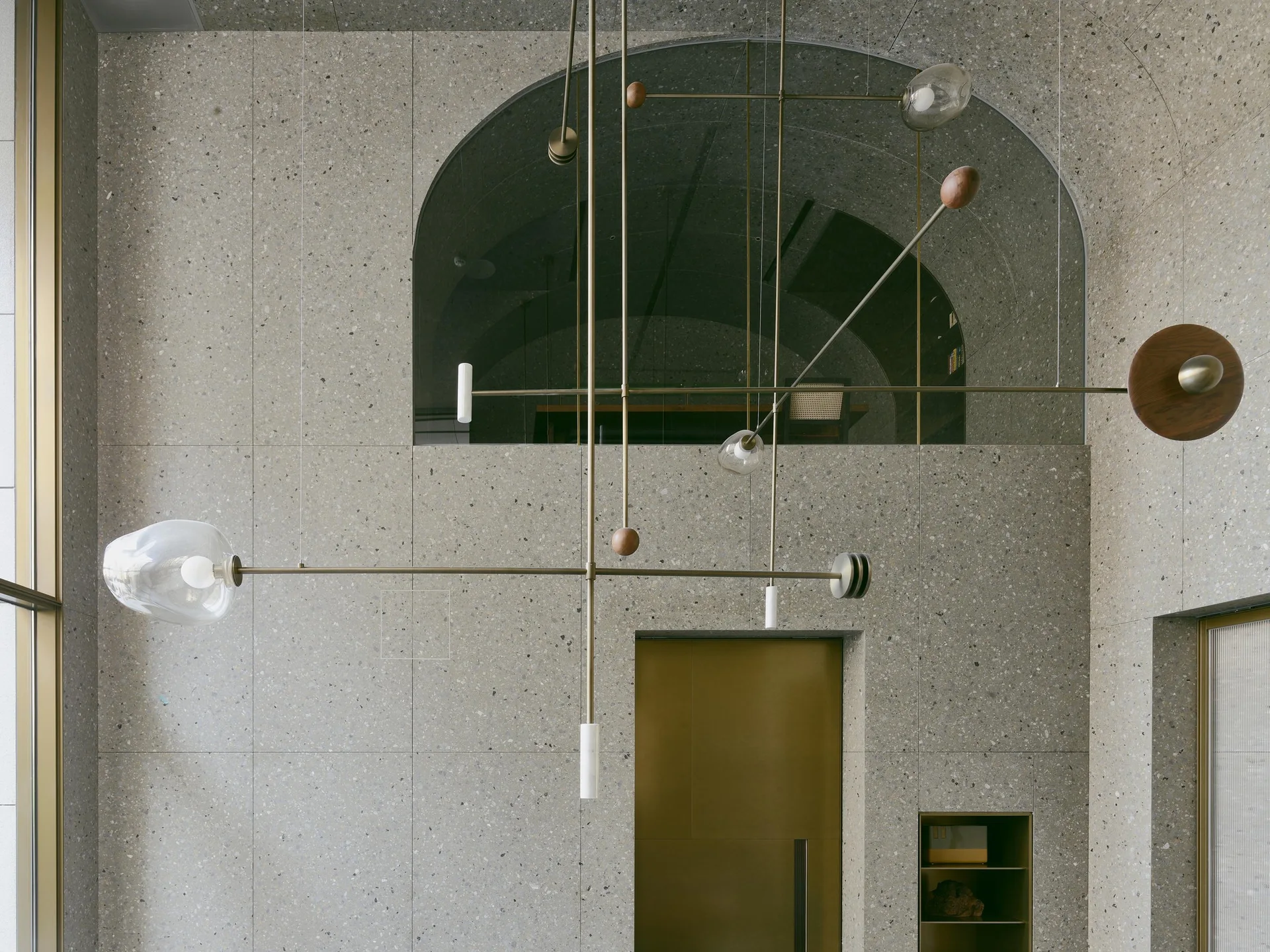 A tall speckled terrazzo wall features a brass door, an upper archway, and a sculptural multi-armed brass pendant light with glass and wood details.