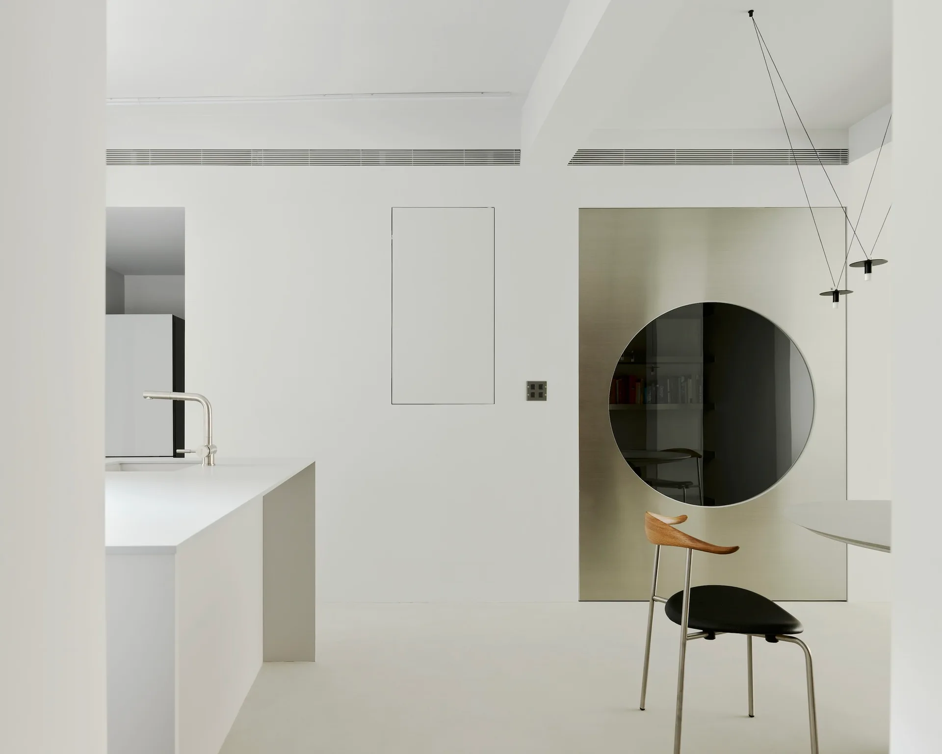 A minimalist white kitchen features a solid white island, a brushed metallic wall panel with a circular cutout window, and a modern dining chair.