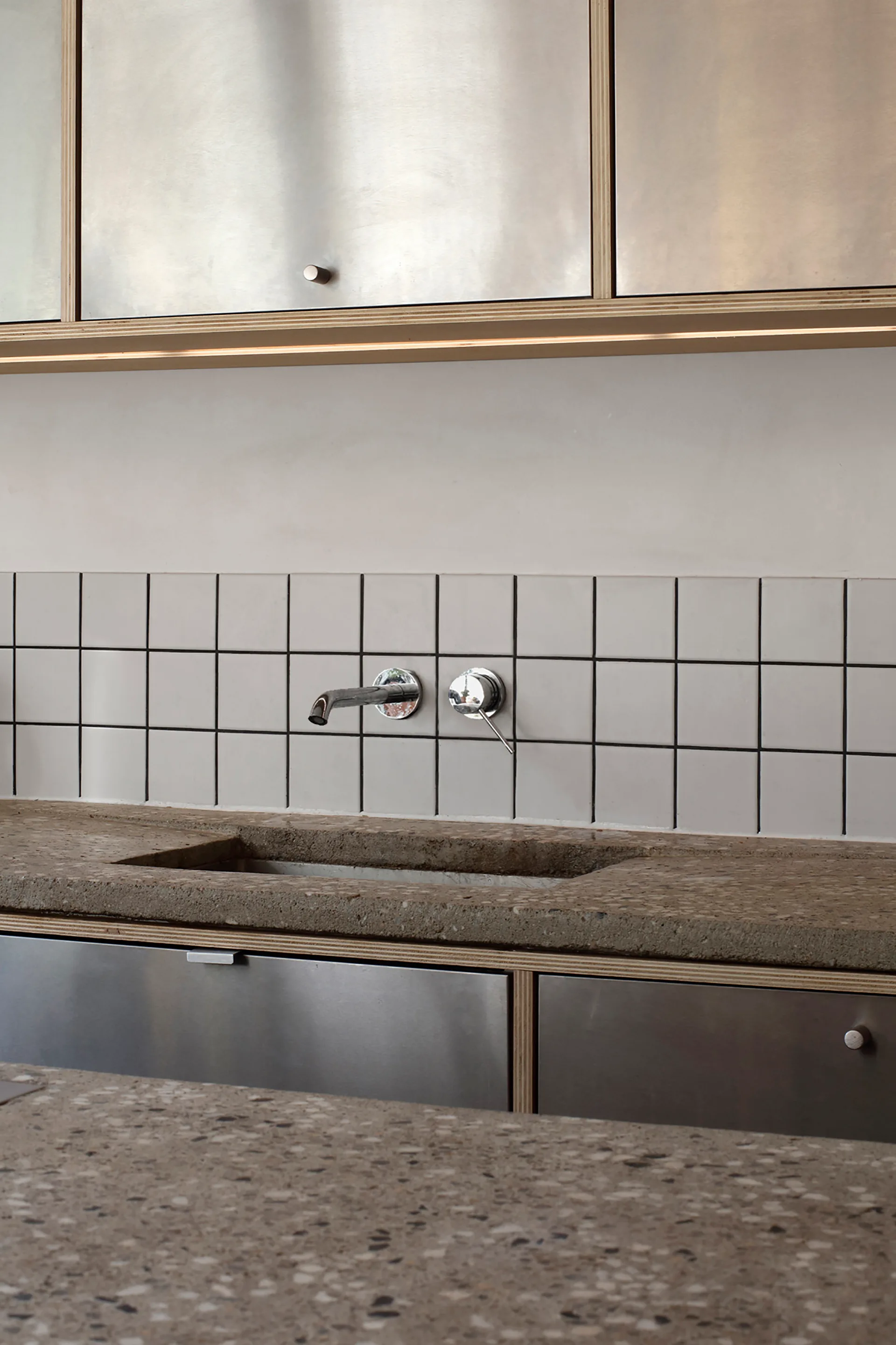 A kitchen counter featuring metal cabinetry with exposed plywood edges, a speckled stone countertop, and a square tile backsplash with a wall-mounted faucet.