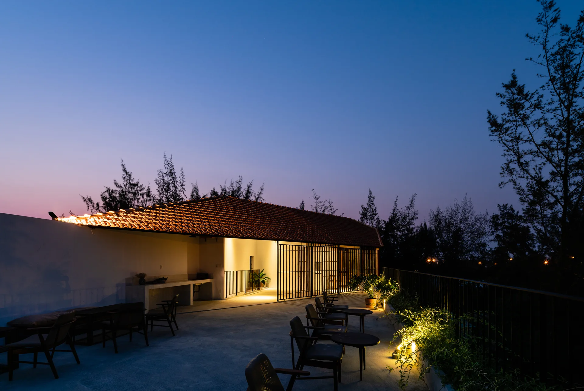 An outdoor concrete terrace features a terracotta tiled roof structure, dark lounge seating, and built-in planters under twilight skies.