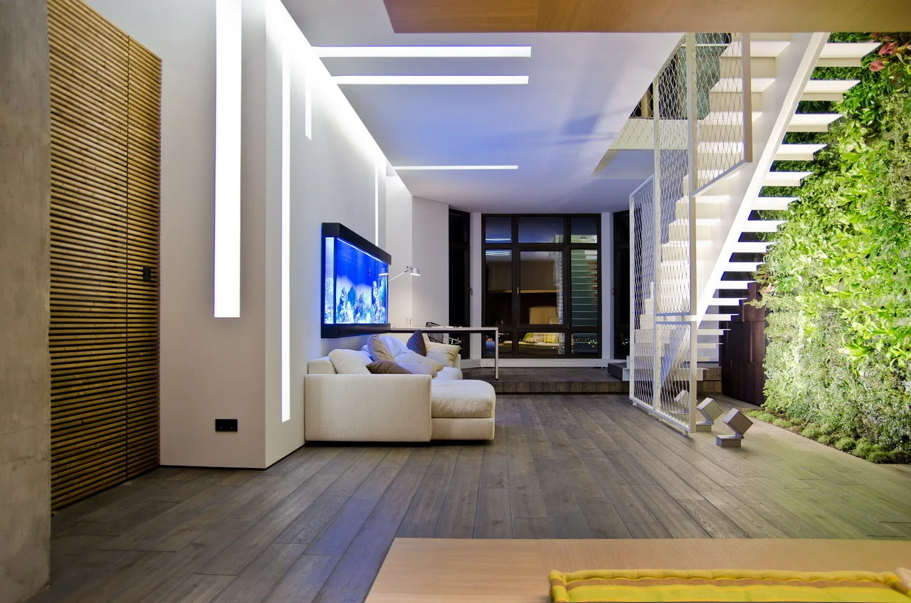 A contemporary living room featuring a white sectional sofa, a wall-mounted blue aquarium, and an open metal staircase next to a lush vertical garden.