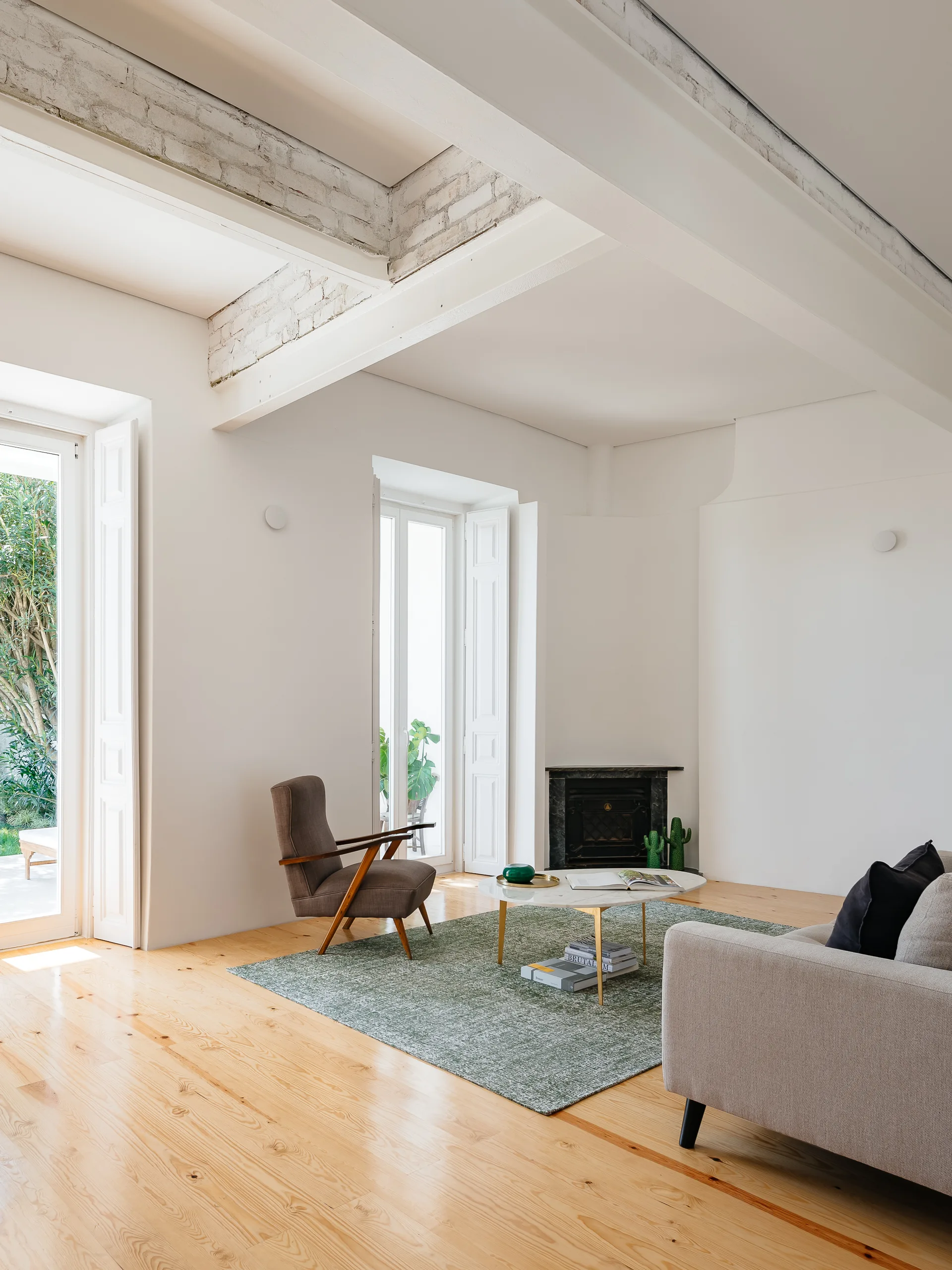 A bright living room features a green rug, a marble-top coffee table, a mid-century armchair, and exposed white ceiling beams over light wood flooring.