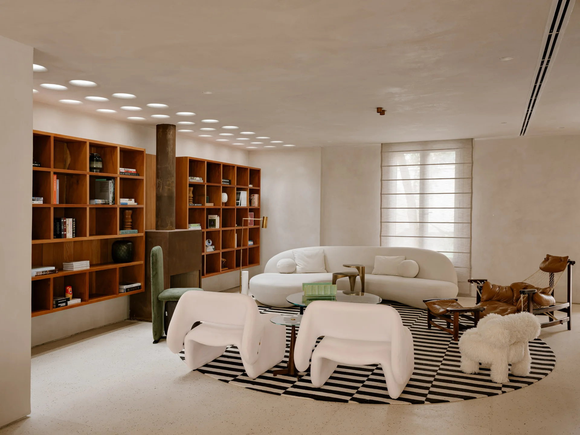 A minimalist living room features a curved white sofa and armchairs arranged on a circular striped rug next to wood built-in bookshelves.