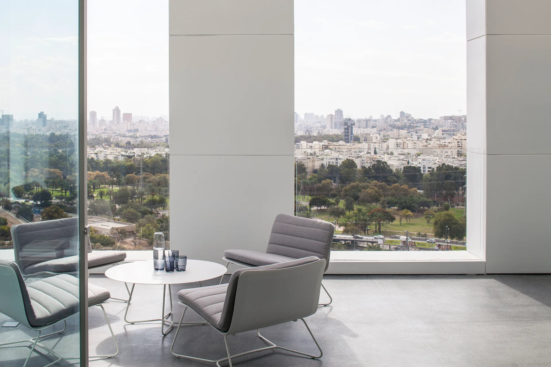 Grey upholstered lounge chairs and a round white table sit on a spacious high-rise terrace framed by white pillars overlooking a panoramic city skyline.