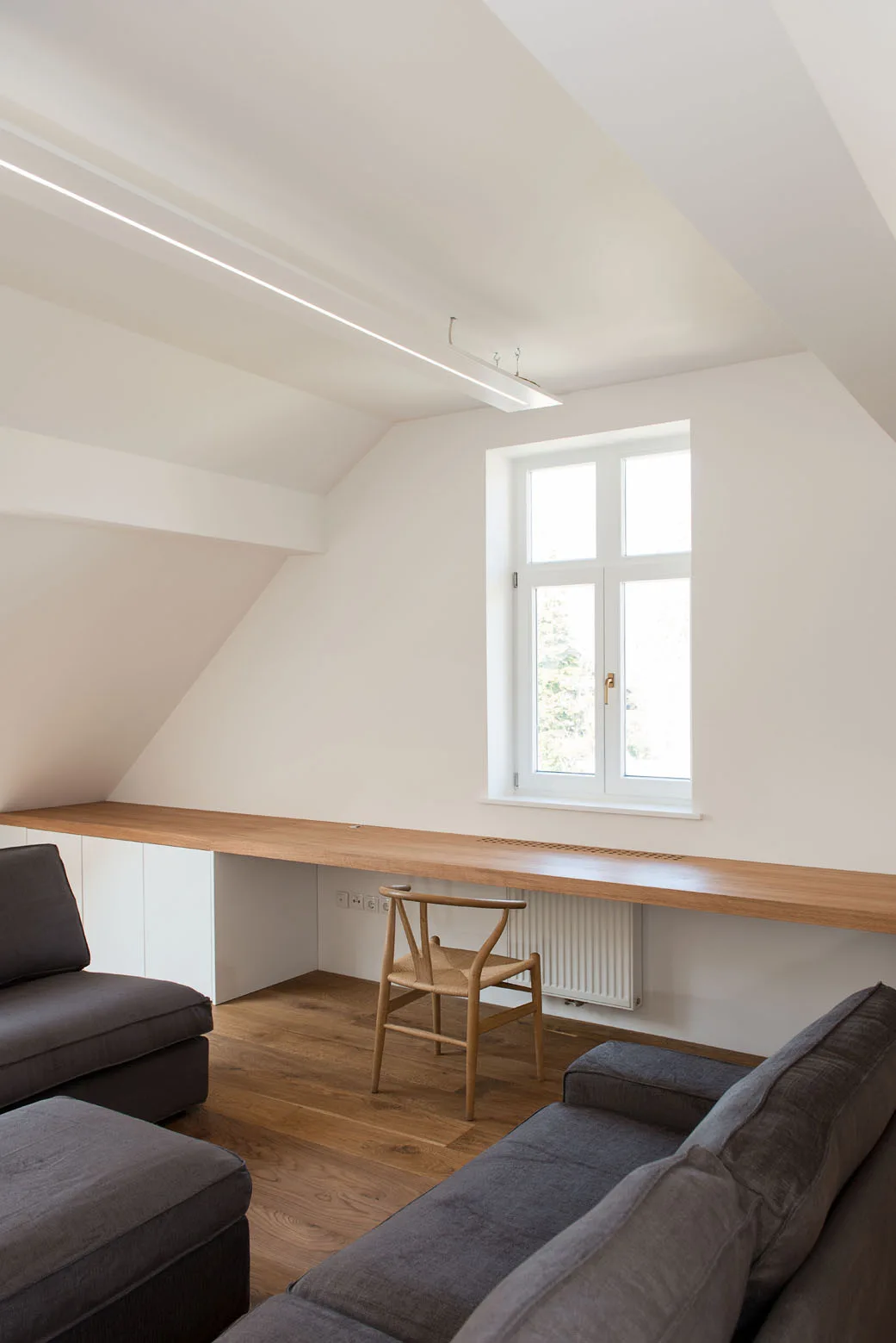 A minimalist attic room features a long wooden desk beneath a window, a classic wishbone chair, and gray fabric seating on wood floors.