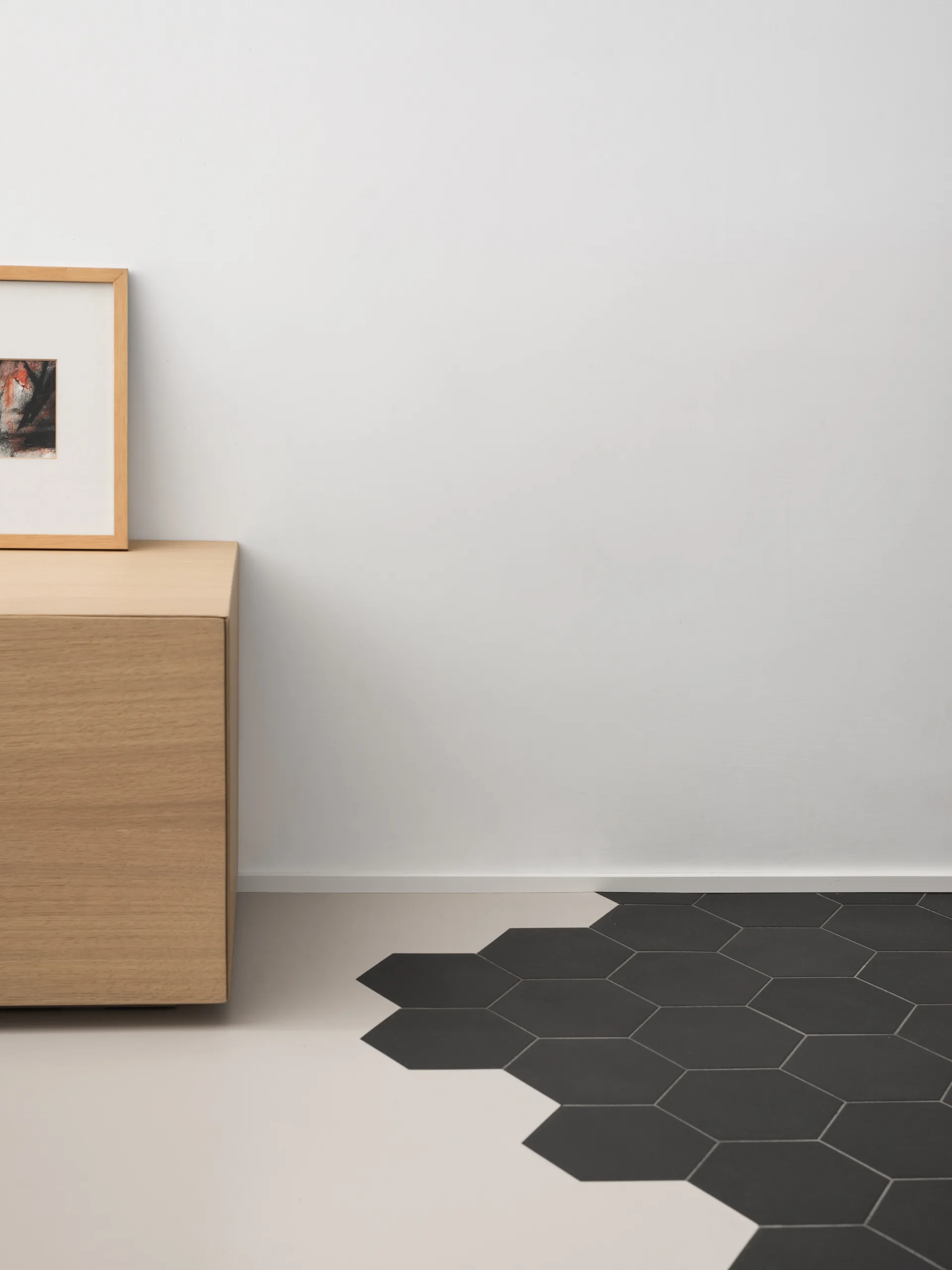 A light wood cabinet stands against a white wall above a floor transition where smooth beige flooring meets black hexagonal tiles.