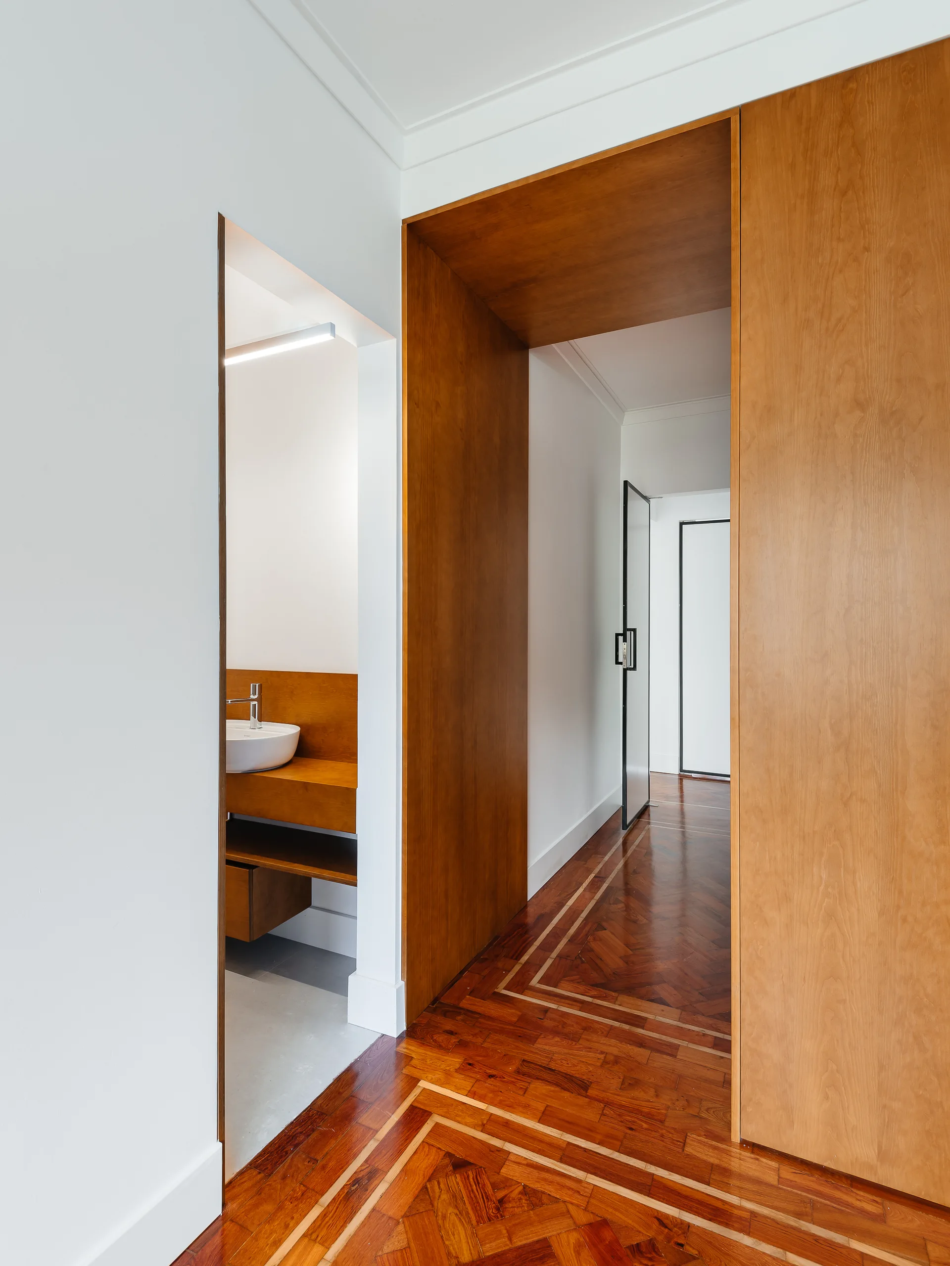 A modern hallway features patterned herringbone parquet flooring, crisp white walls, a large built-in wood portal, and a glimpse into a powder room.