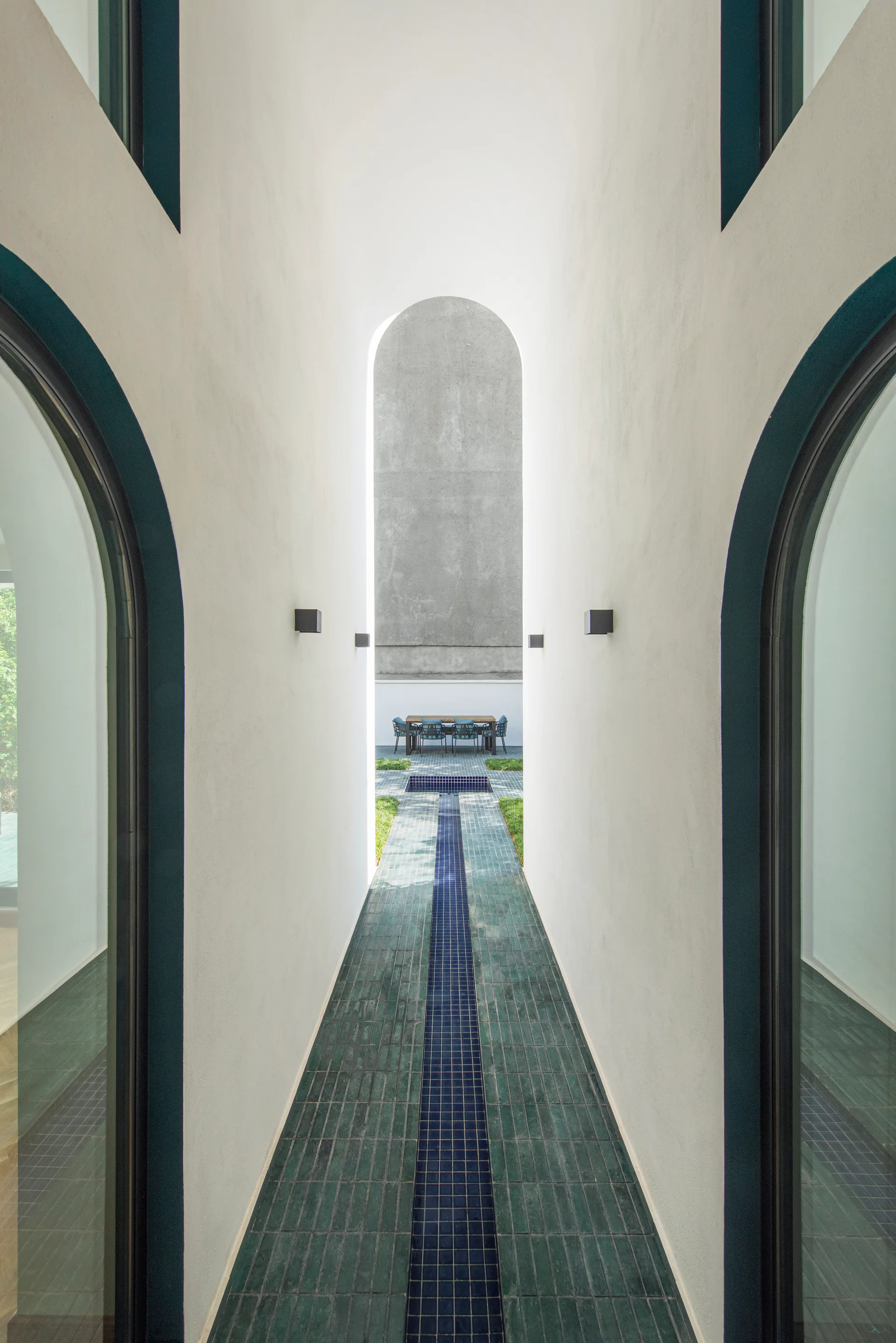 A long white arched outdoor passage with a glossy green tiled walkway and a central blue mosaic strip leading to a distant dining table.