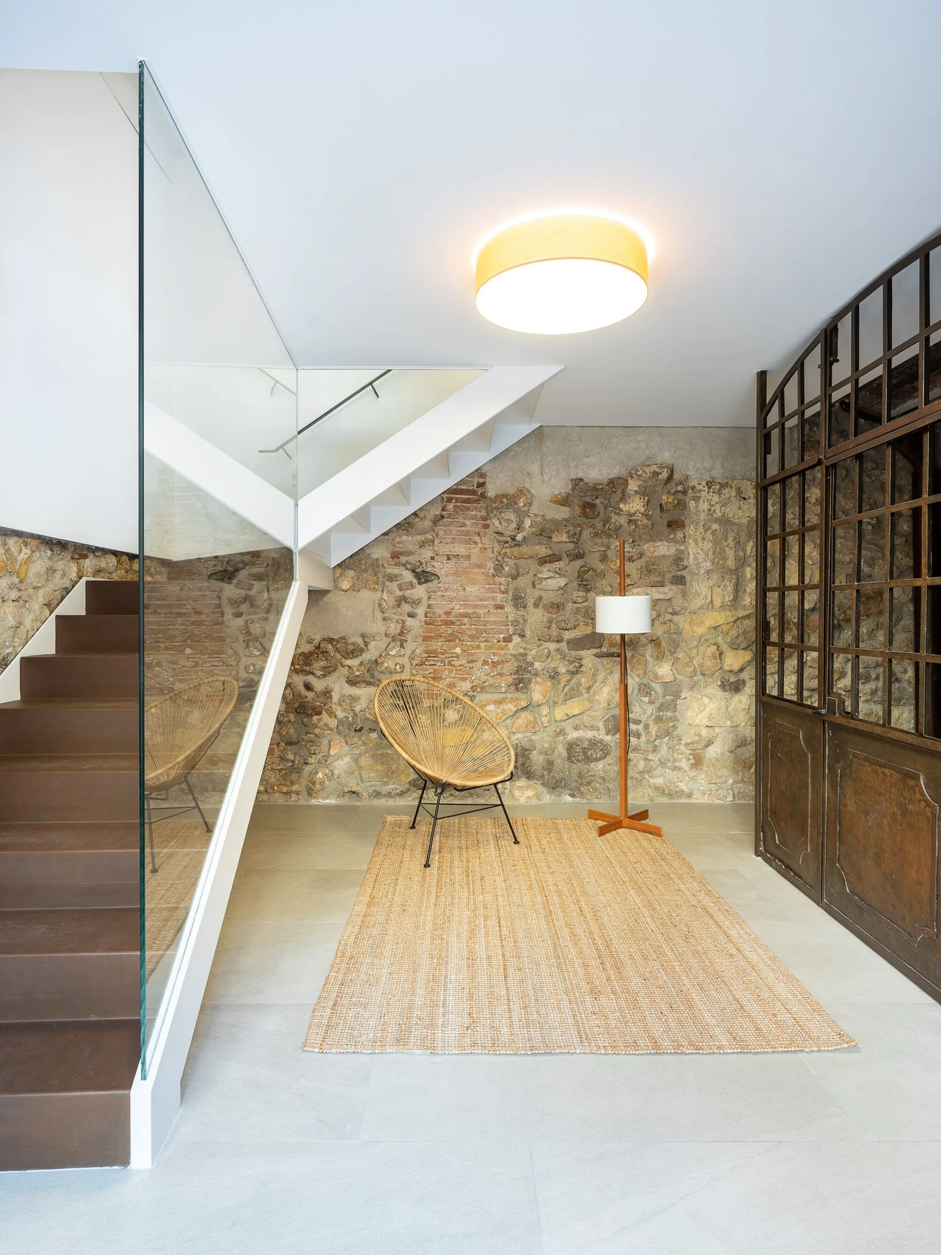 A modern staircase with a glass railing stands next to an exposed stone wall, featuring a woven accent chair, floor lamp, and a jute rug.
