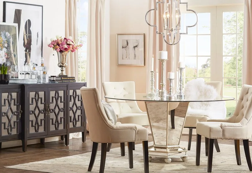 A dining room features a round glass-top table with a mirrored base, cream tufted chairs, and a dark wood sideboard with geometric fretwork doors.