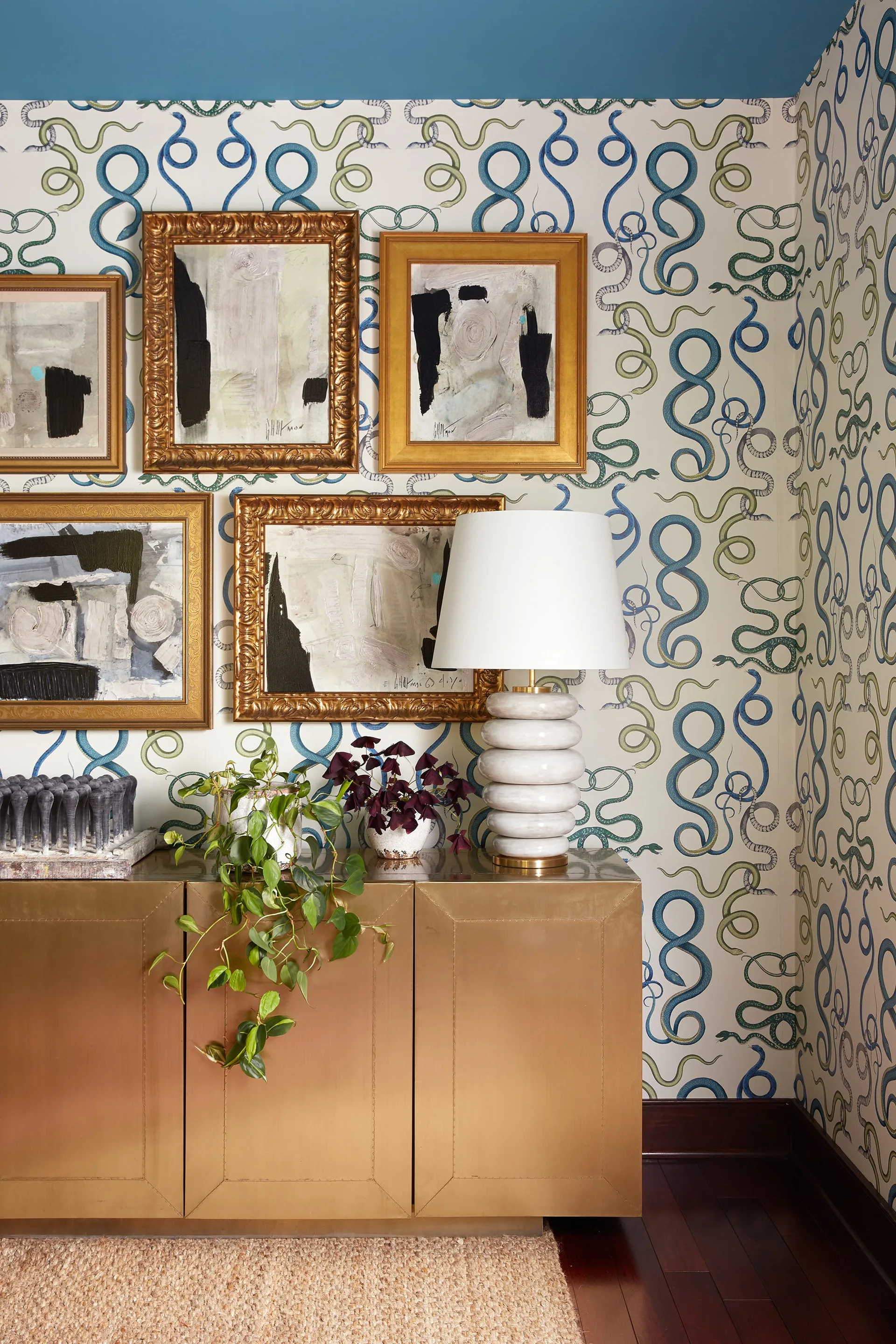 A brass credenza stands against blue snake-patterned wallpaper below a gallery wall of abstract art in gold frames.