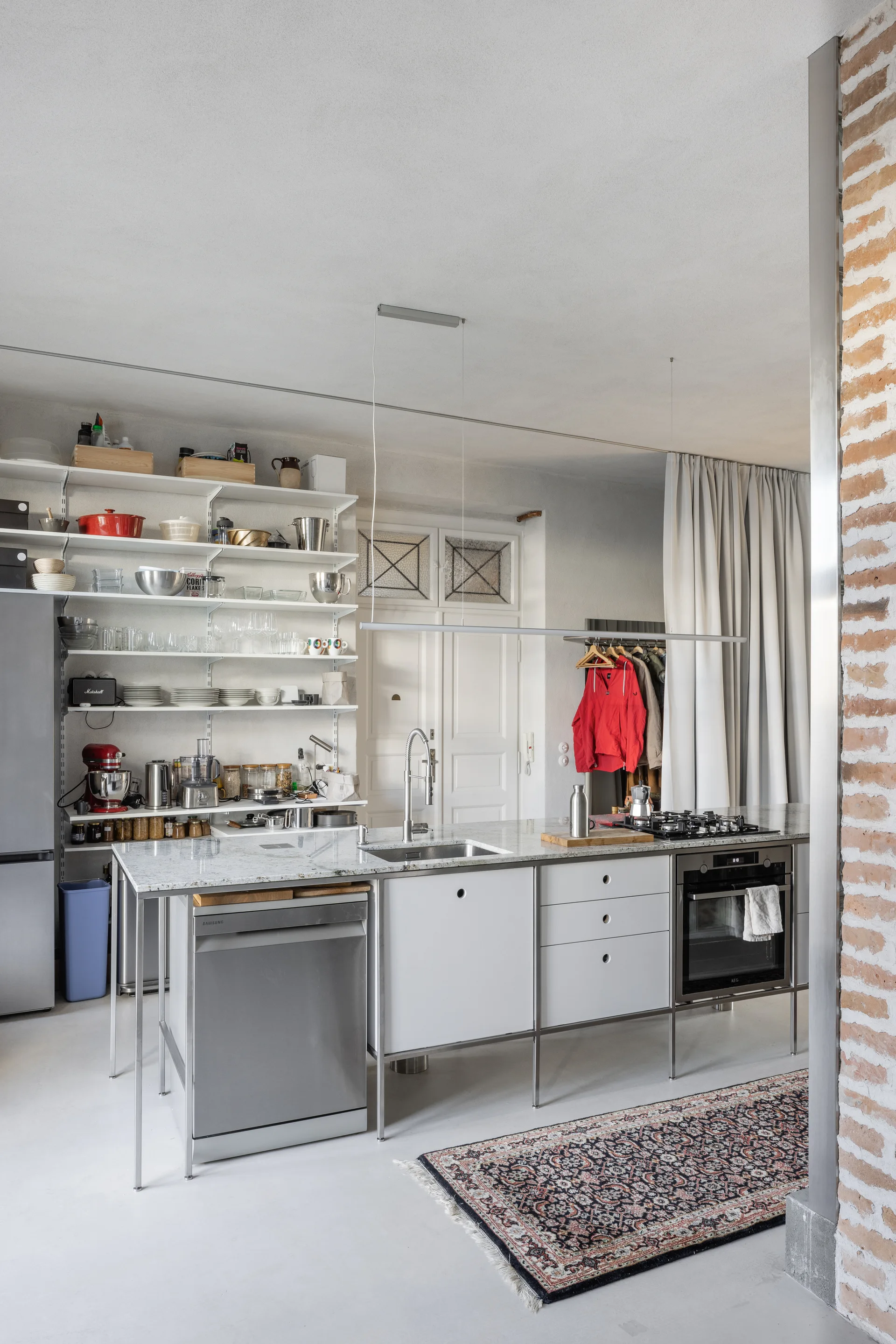 A modern kitchen with a stainless steel island, integrated sink and cooktop, open metal wall shelves, and an exposed brick column.