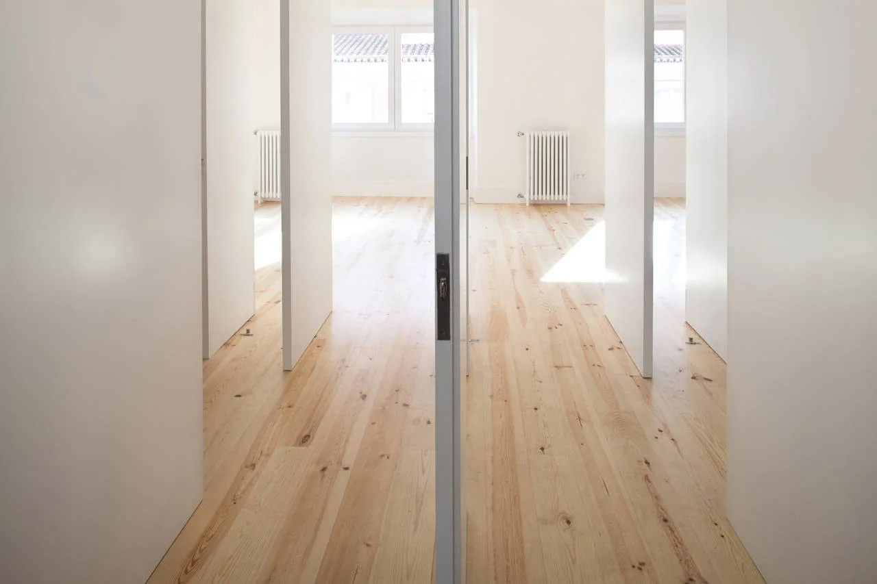 A bright minimalist hallway features light wood plank floors, smooth white walls, and open frameless white doors leading to adjacent rooms with radiators.