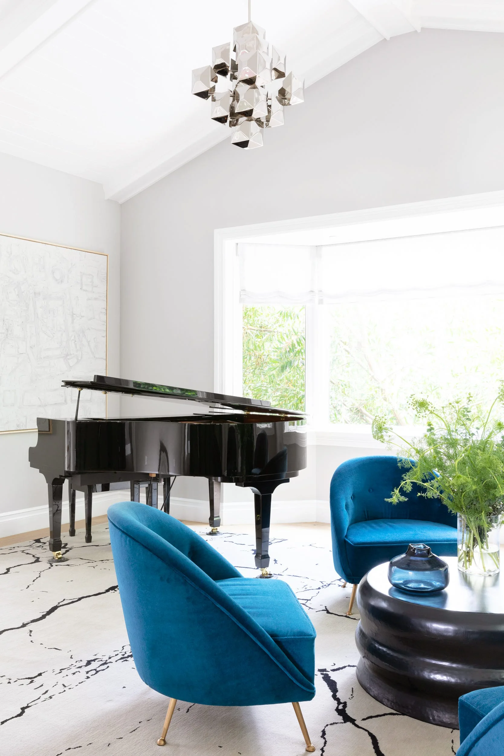 A light gray living room features a black grand piano, blue velvet armchairs, a geometric silver chandelier, and a large abstract patterned rug.