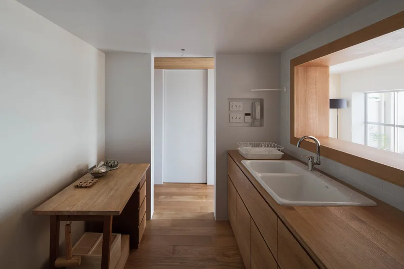 A minimalist kitchen featuring natural oak cabinetry, matching countertops with an integrated white sink, and a wooden pass-through window.