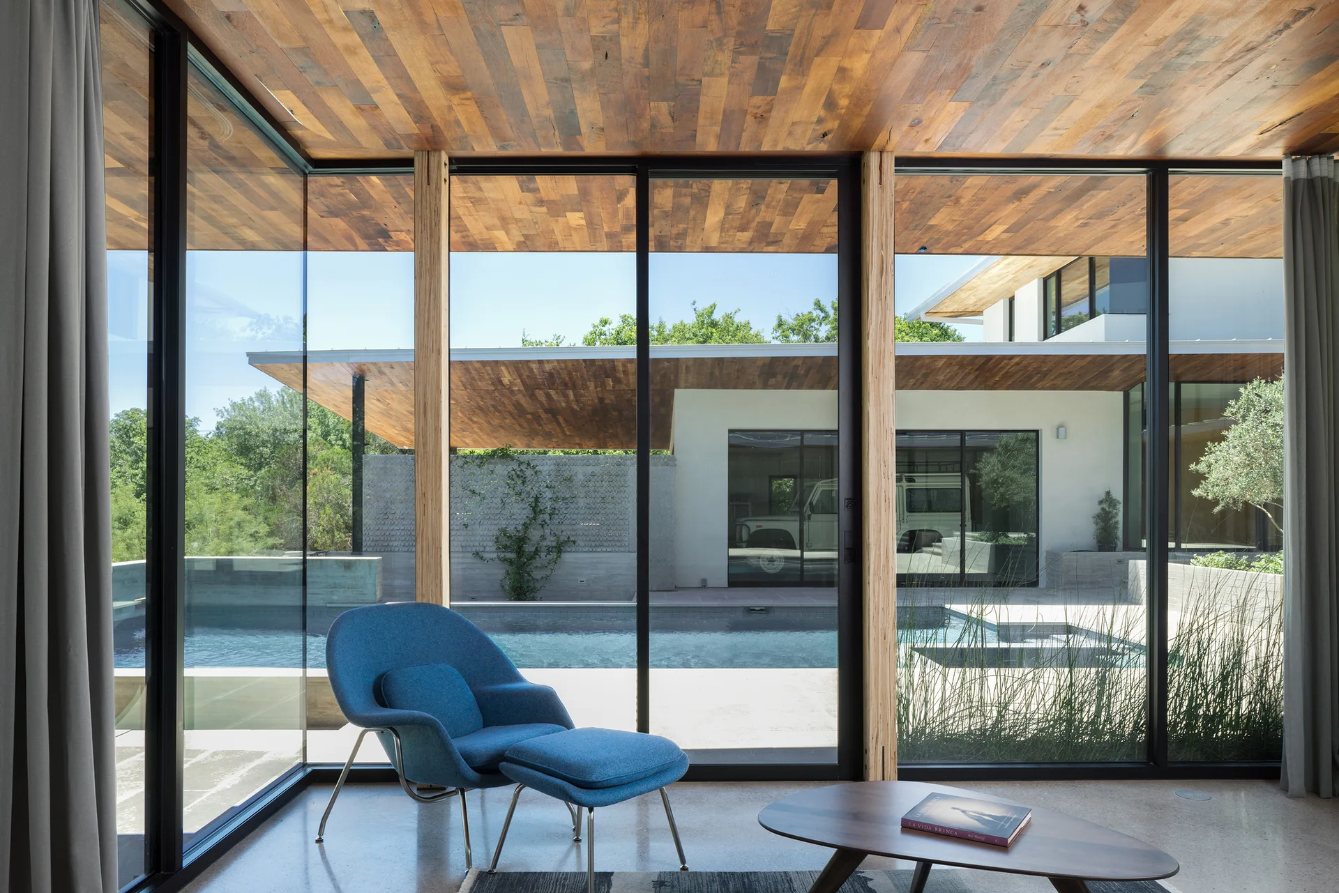 A blue lounge chair with matching ottoman sits in a living room featuring a warm wood ceiling and floor-to-ceiling glass doors facing an outdoor pool.