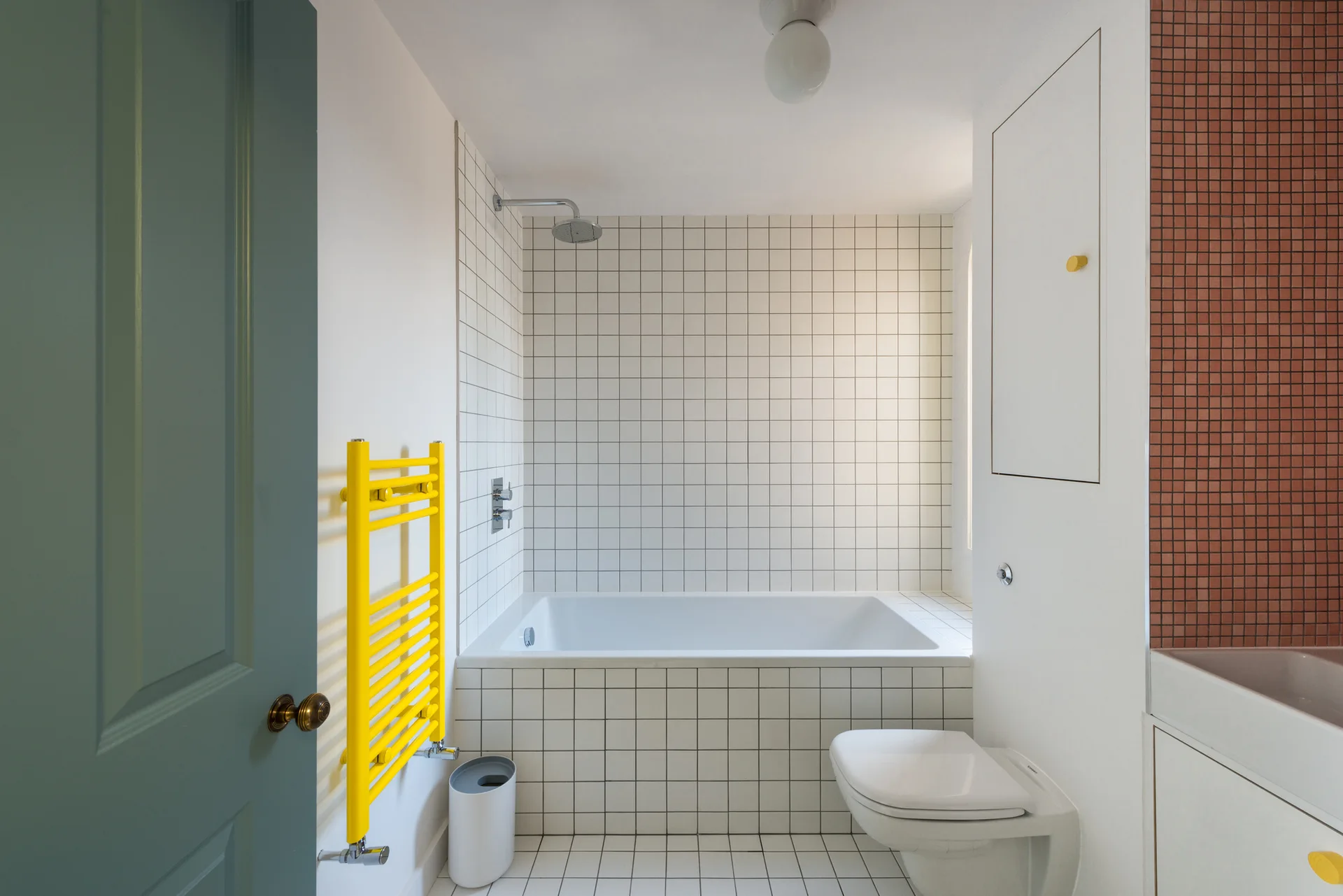 A minimalist modern bathroom features a white tiled bathtub, white grid wall tiles, and a bright yellow towel radiator.