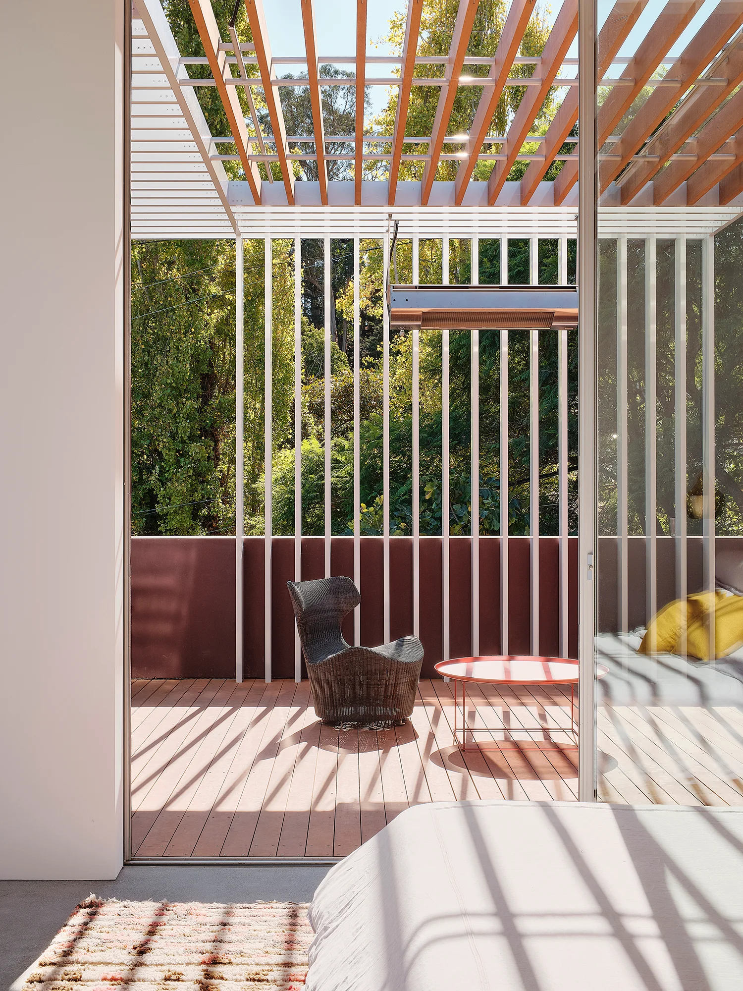 A modern wooden balcony viewed from an interior bedroom featuring a timber pergola, vertical white railing slats, and a dark wicker armchair.