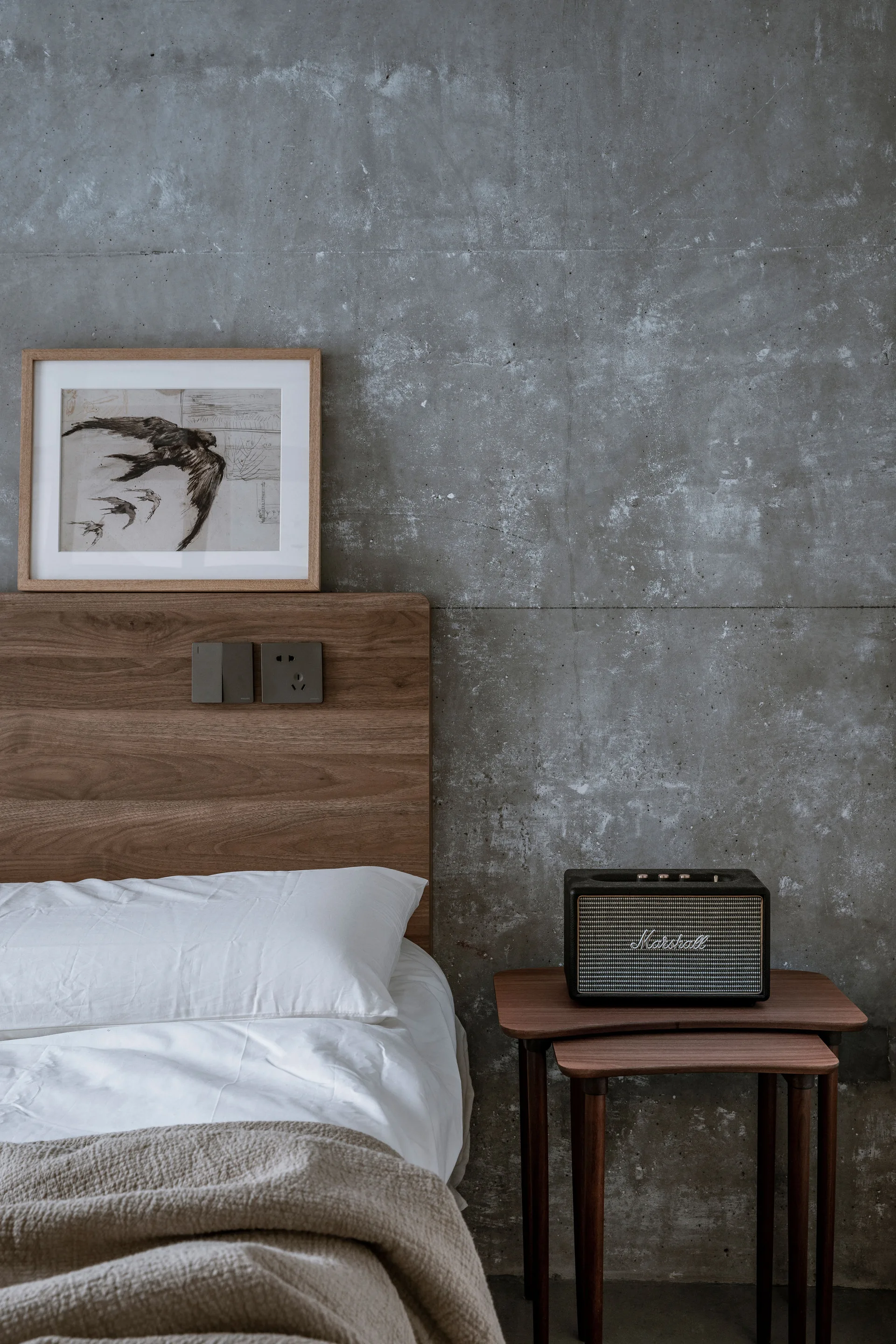 A bedroom corner features a wooden headboard against a raw concrete wall, a white pillow, and a retro speaker on a wooden nesting side table.