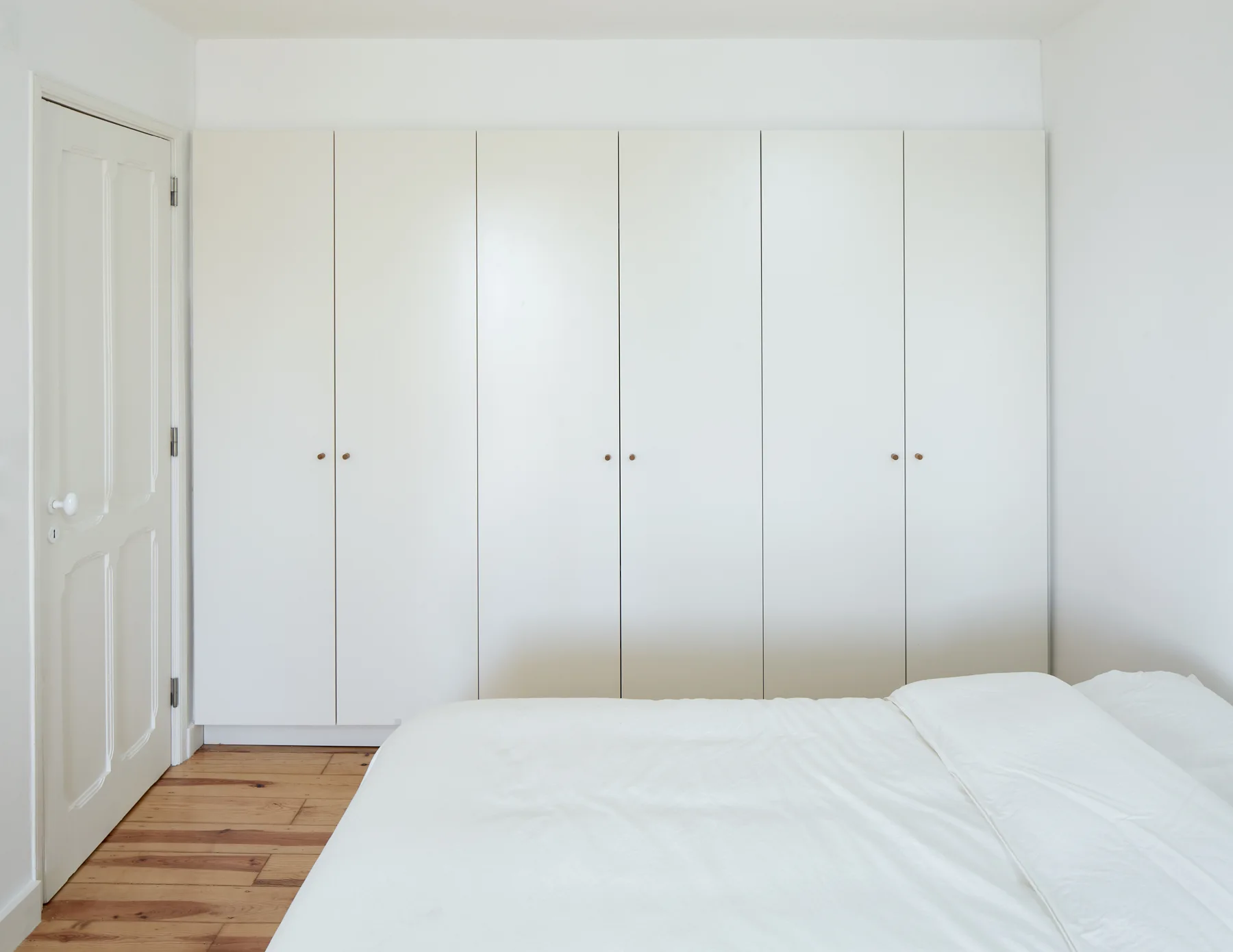 A minimalist white bedroom features a floor-to-ceiling built-in wardrobe and a white bed on natural pine wood flooring.