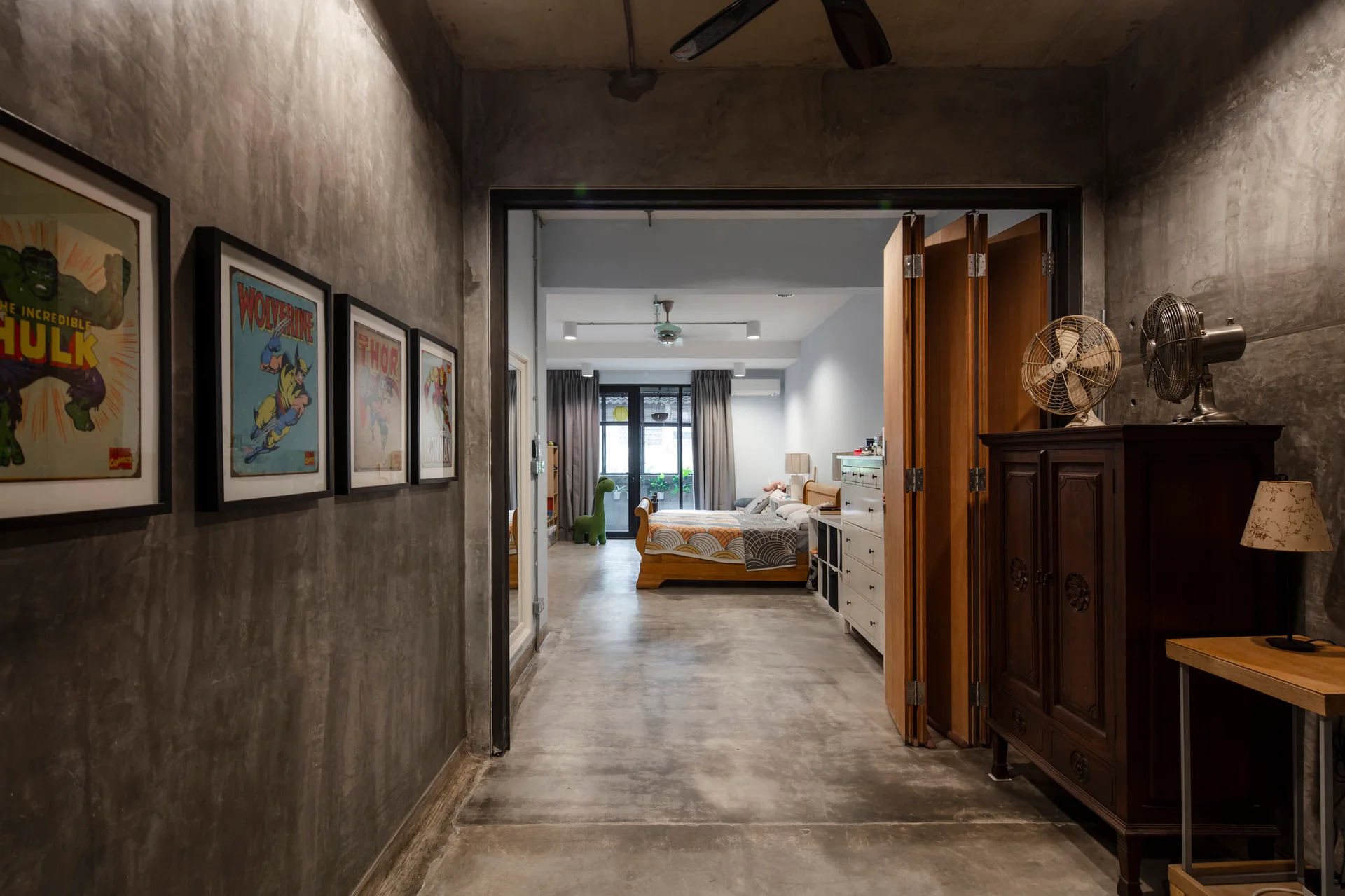 A view through a doorway into a bedroom featuring raw concrete walls, a wooden folding partition, and framed superhero prints in the hallway.