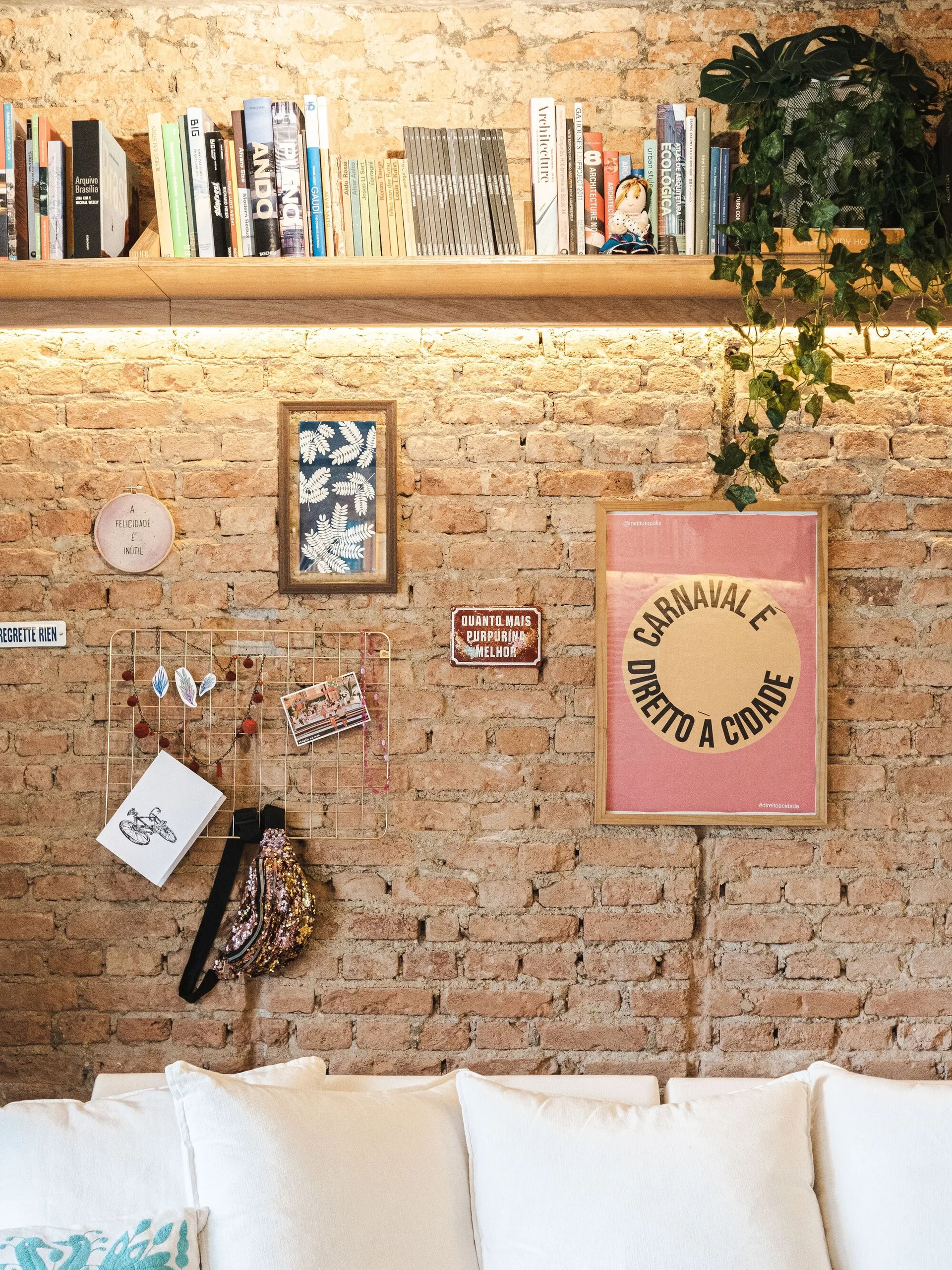 An exposed brick wall features a wooden bookshelf with under-shelf lighting, eclectic framed art prints, and white sofa cushions below.