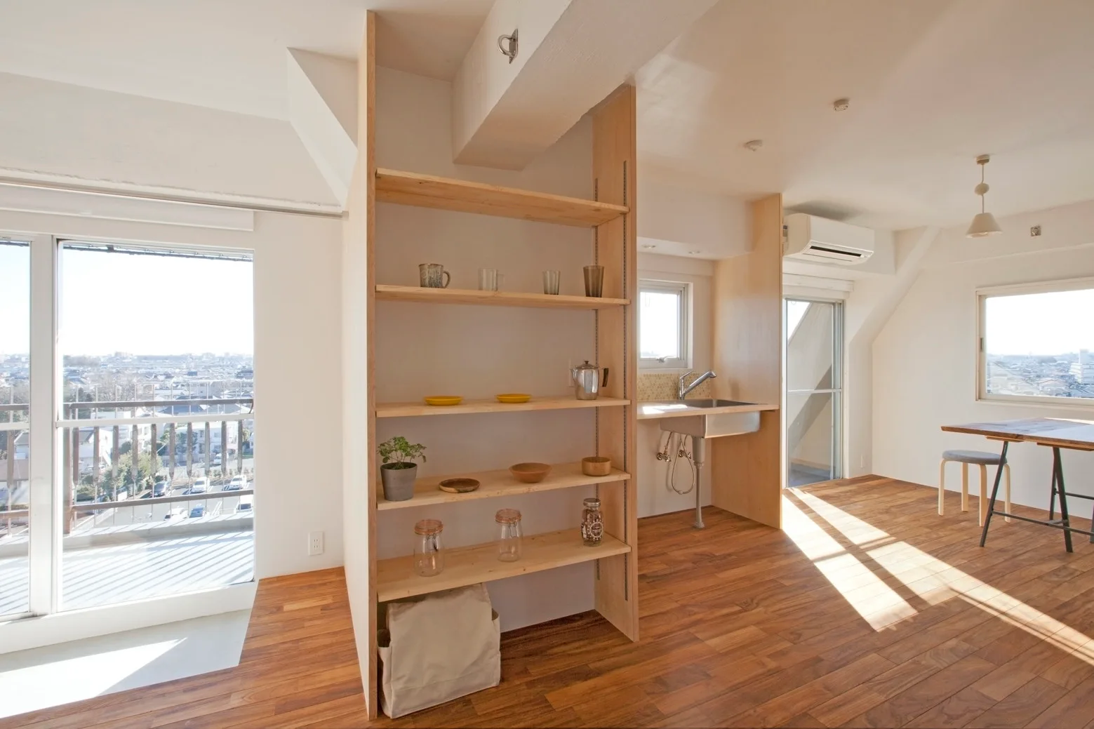 A tall light wood open shelving unit acts as a room divider in a bright open-plan kitchen and living space with warm wood floors.