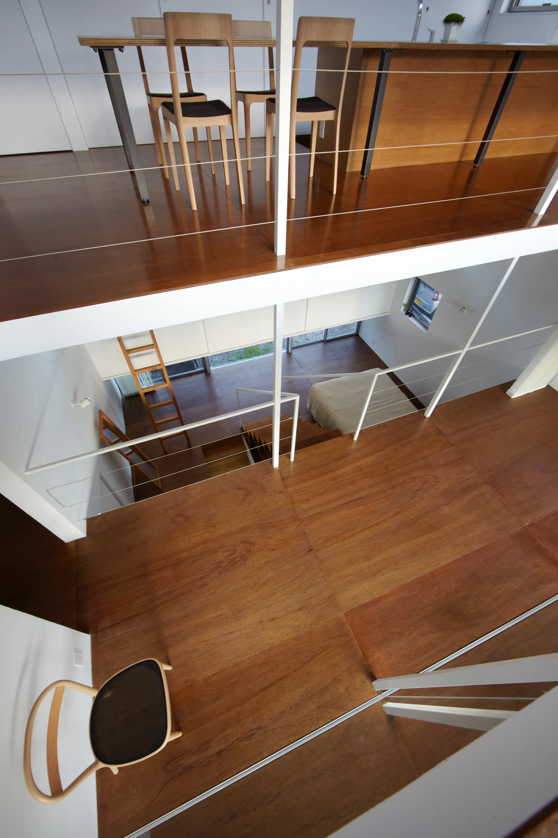 High-angle view of a split-level modern home featuring warm wooden flooring, a mezzanine dining area with wooden chairs, and a lower-level bedroom.