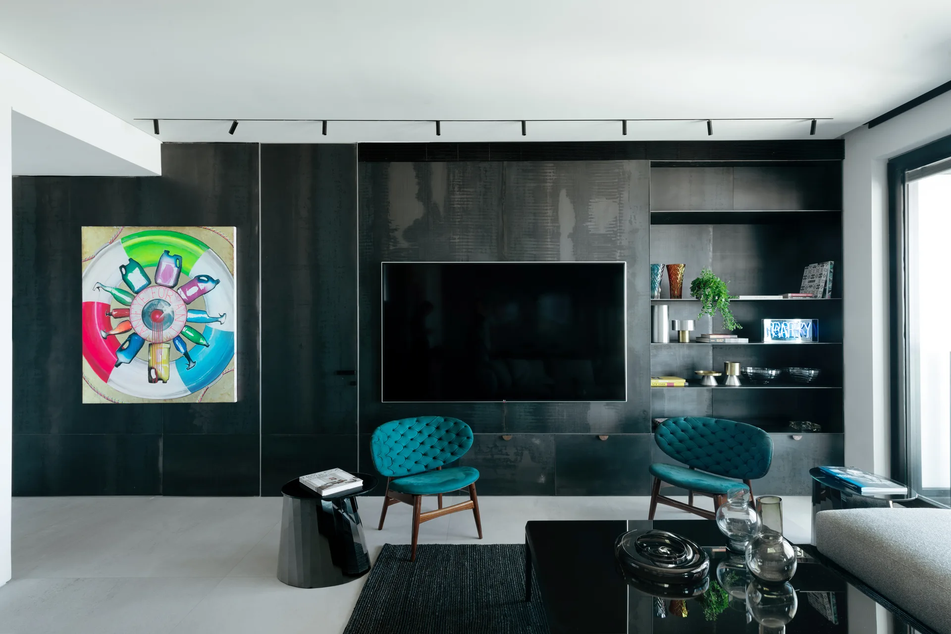 A contemporary living room features a dark metal paneled wall with a mounted TV, built-in shelves, and two teal tufted armchairs.
