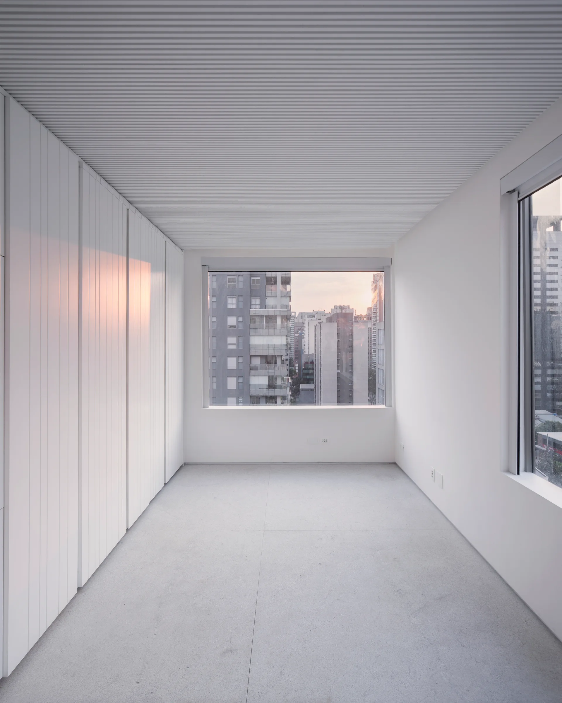 An empty white room with a textured slatted ceiling, a wall of tall built-in closets, and a large window looking out at high-rise buildings.