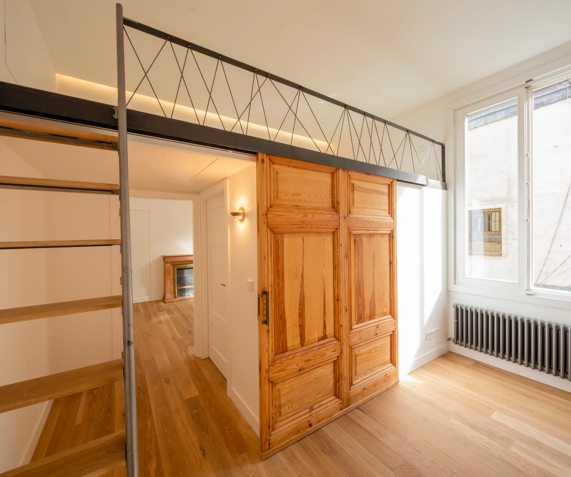 A bright residential room features a paneled wood sliding door, a metal loft ladder, and light wood flooring next to a large window.