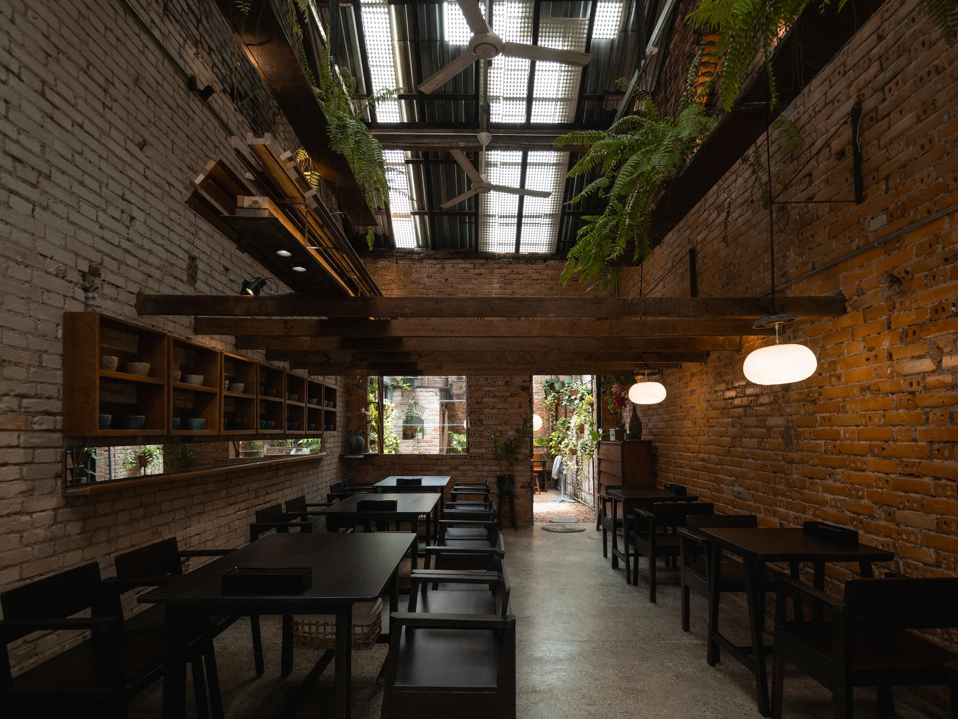 A spacious dining area featuring black wooden tables and chairs set between exposed brick walls beneath a skylight roof with hanging ferns.